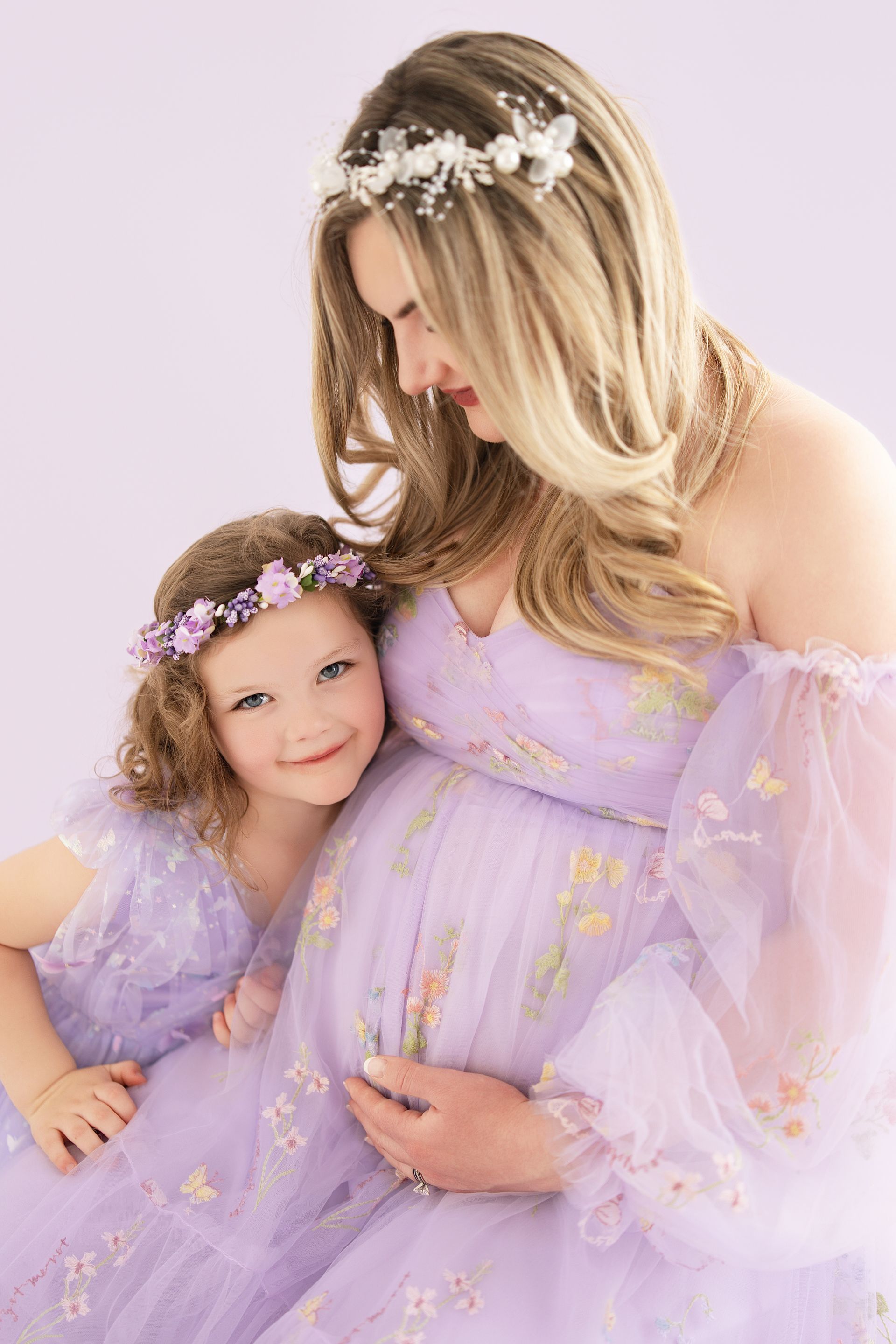 Mother showing her baby bump while daughter smiles in a purple mother-daughter studio session