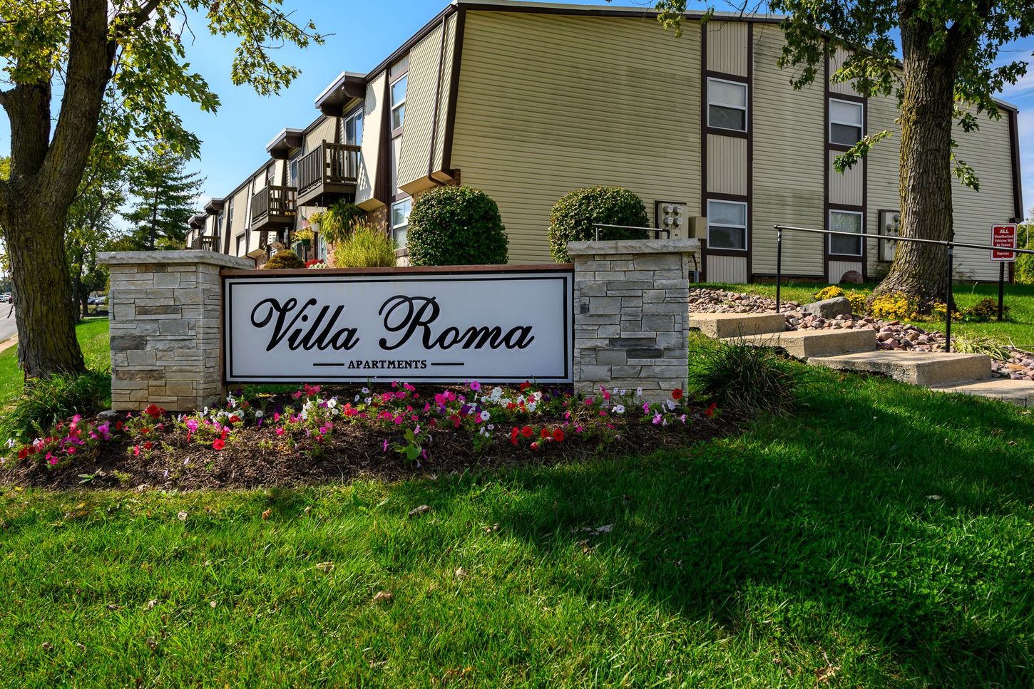 Villa Roma apartment complex sign with building in the background. Green grass and trees frame the scene.