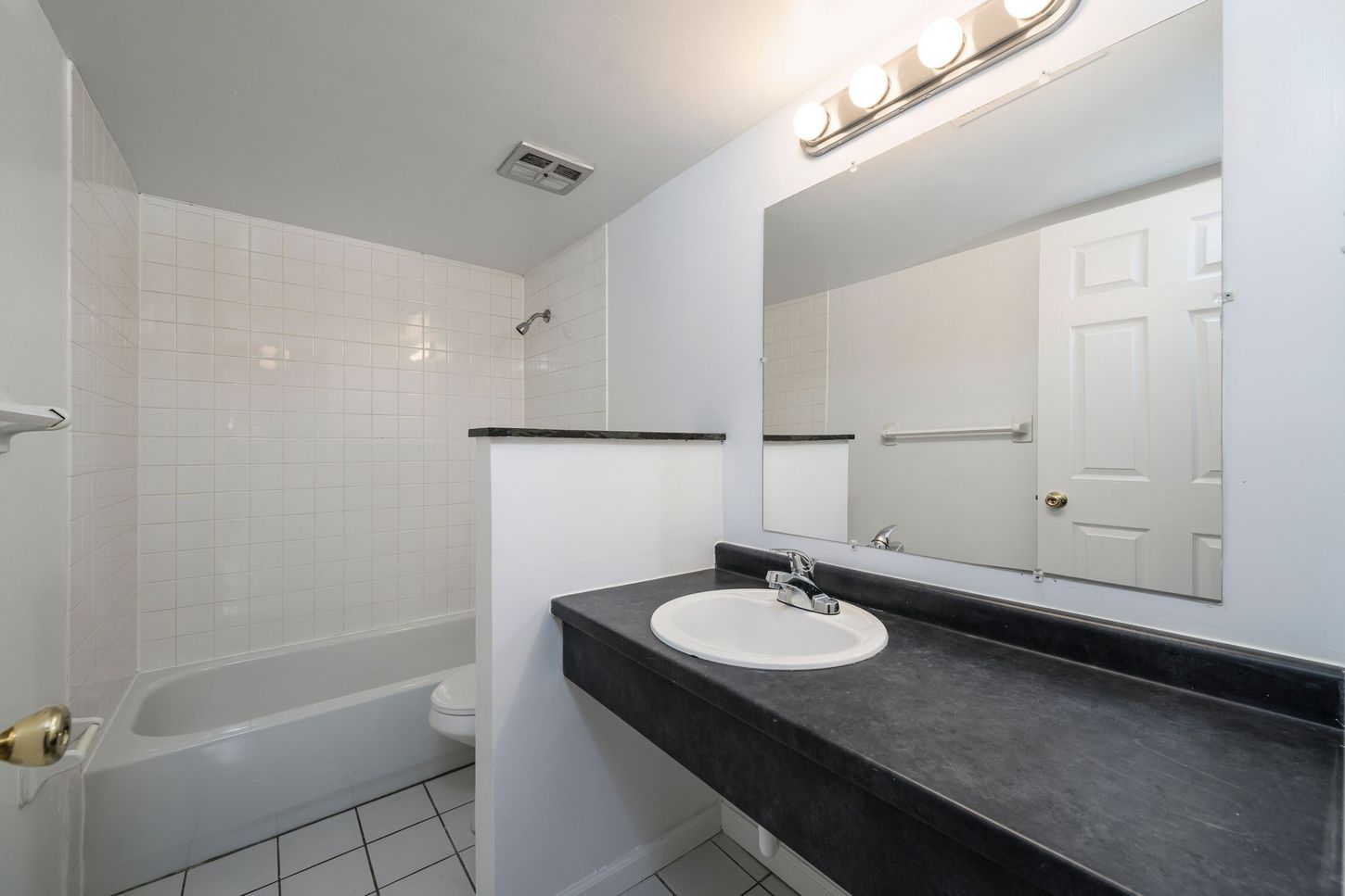 Bathroom with white tile, dark countertop, large mirror, and a white door.