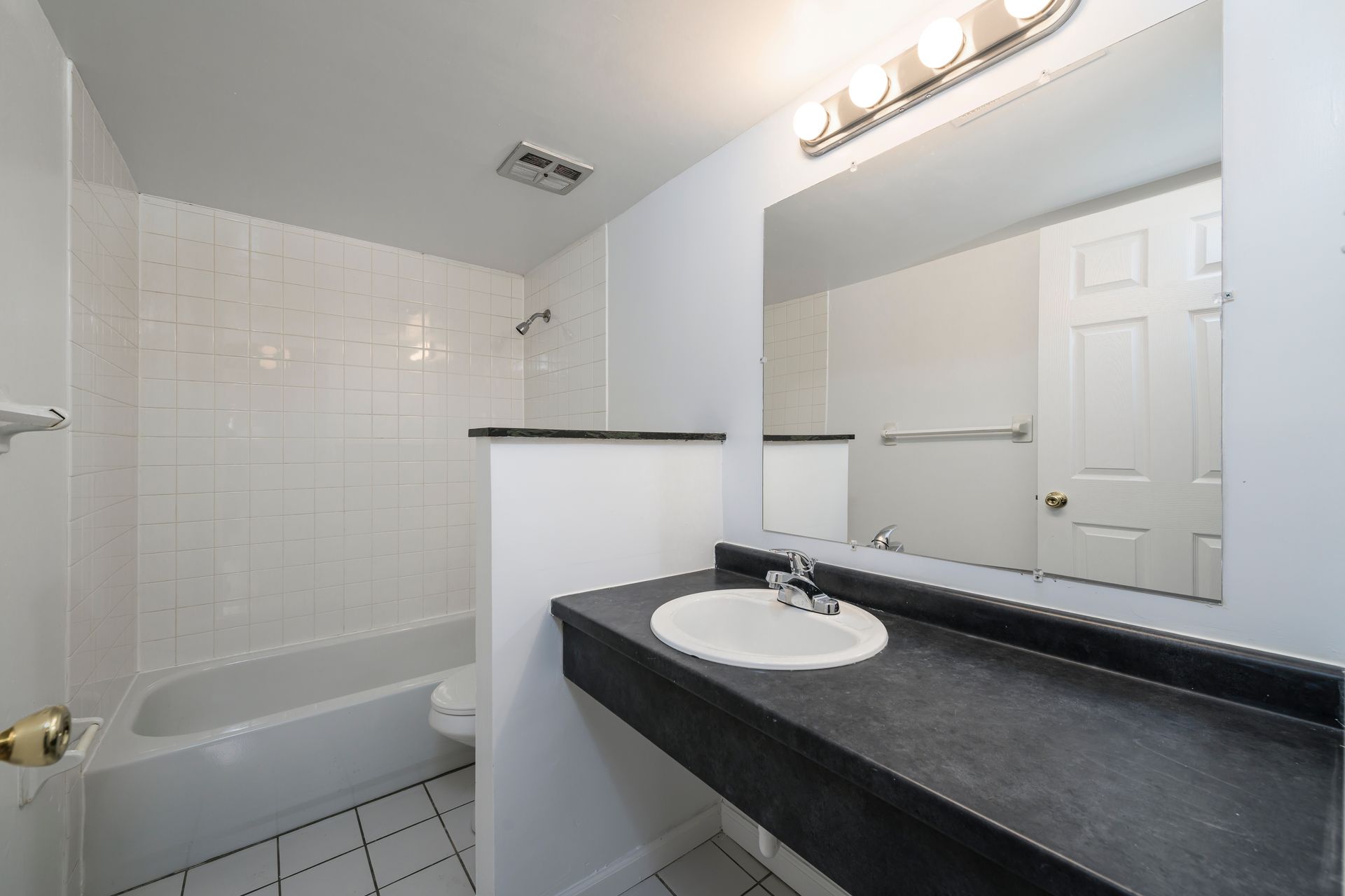 Bathroom with white tile, dark countertop, large mirror, and a white door.
