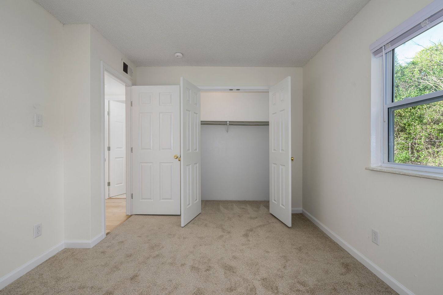 Empty bedroom with closet, window, and door, beige carpet and walls.