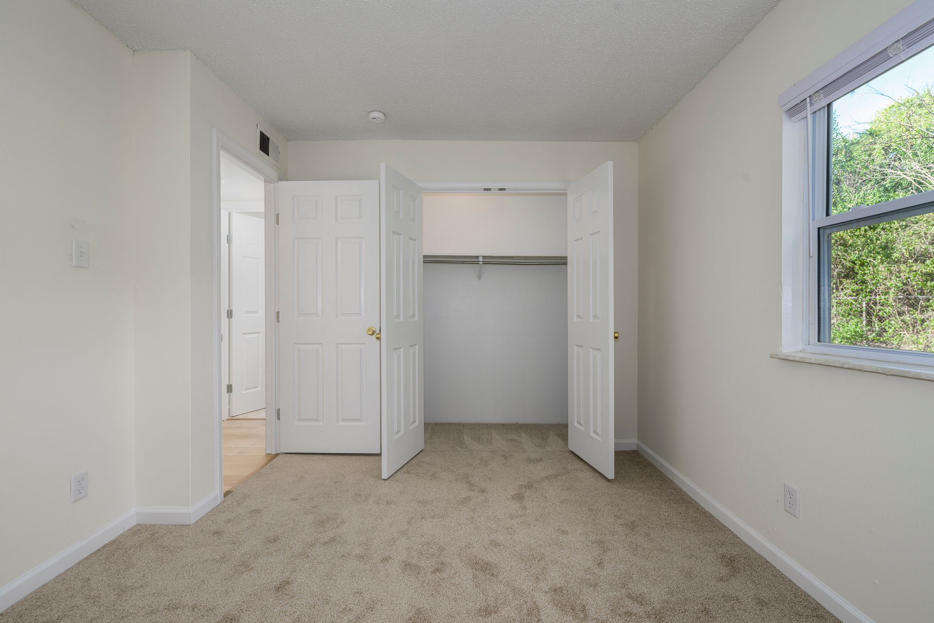 Empty bedroom with closet, window, and door, beige carpet and walls.