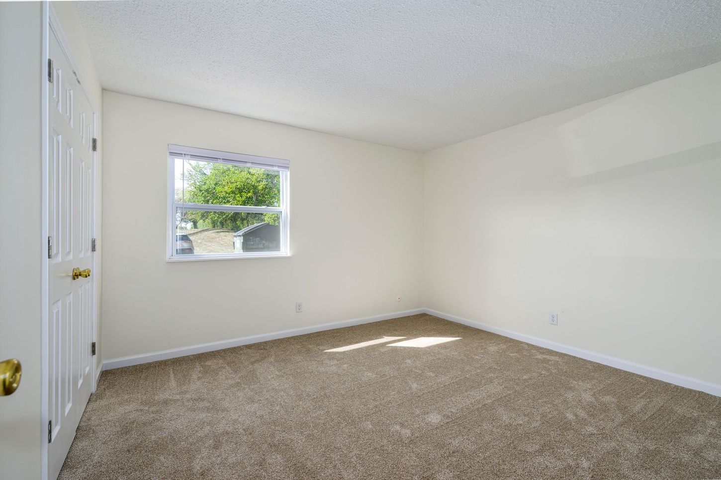 Empty bedroom with beige carpet, white walls, small window, and closet doors.