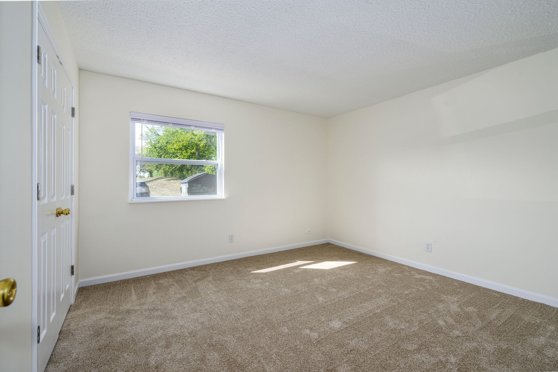 Empty bedroom with beige carpet, white walls, small window, and closet doors.