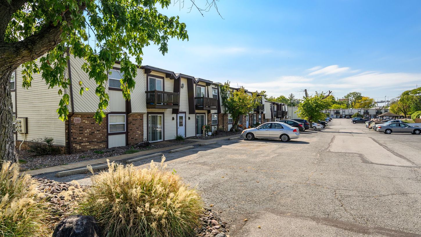 Apartment complex with parking lot, cars, and tree in sunny setting.