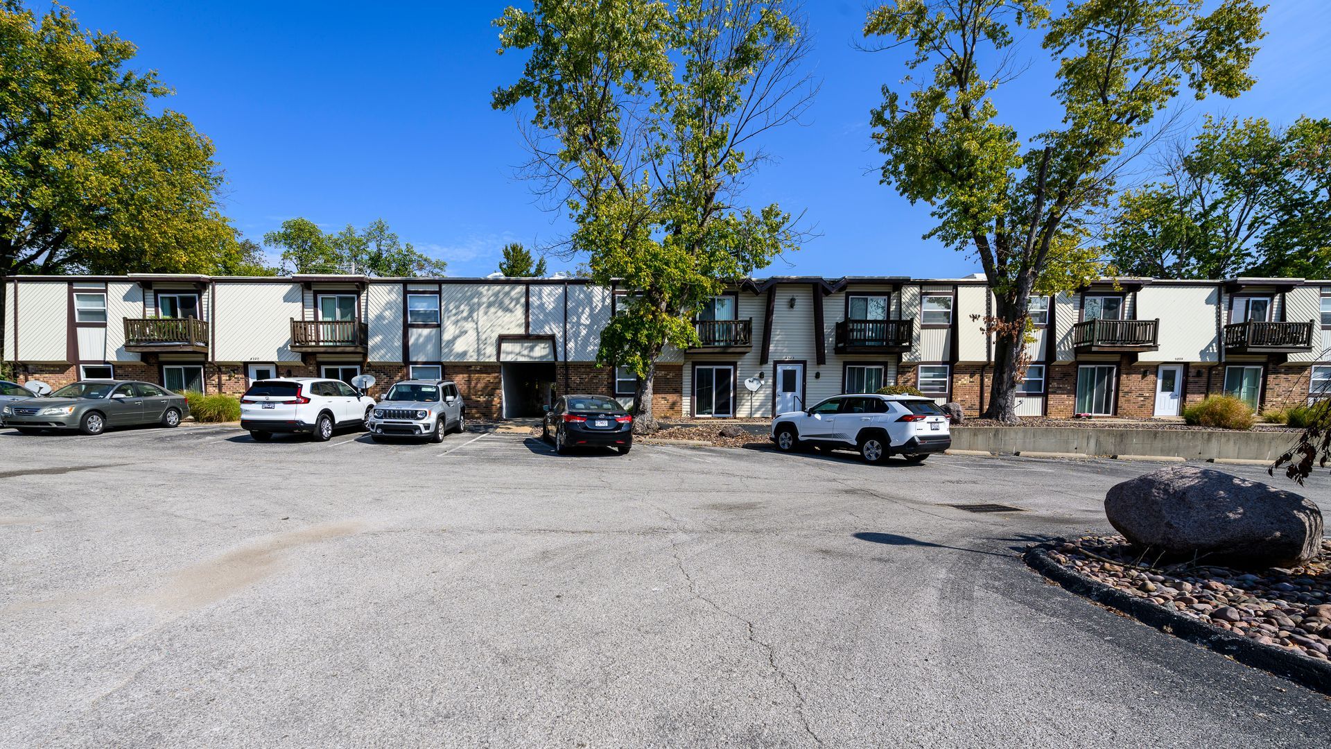 Apartment building exterior with parked cars in a gravel parking lot. Trees and blue sky visible.