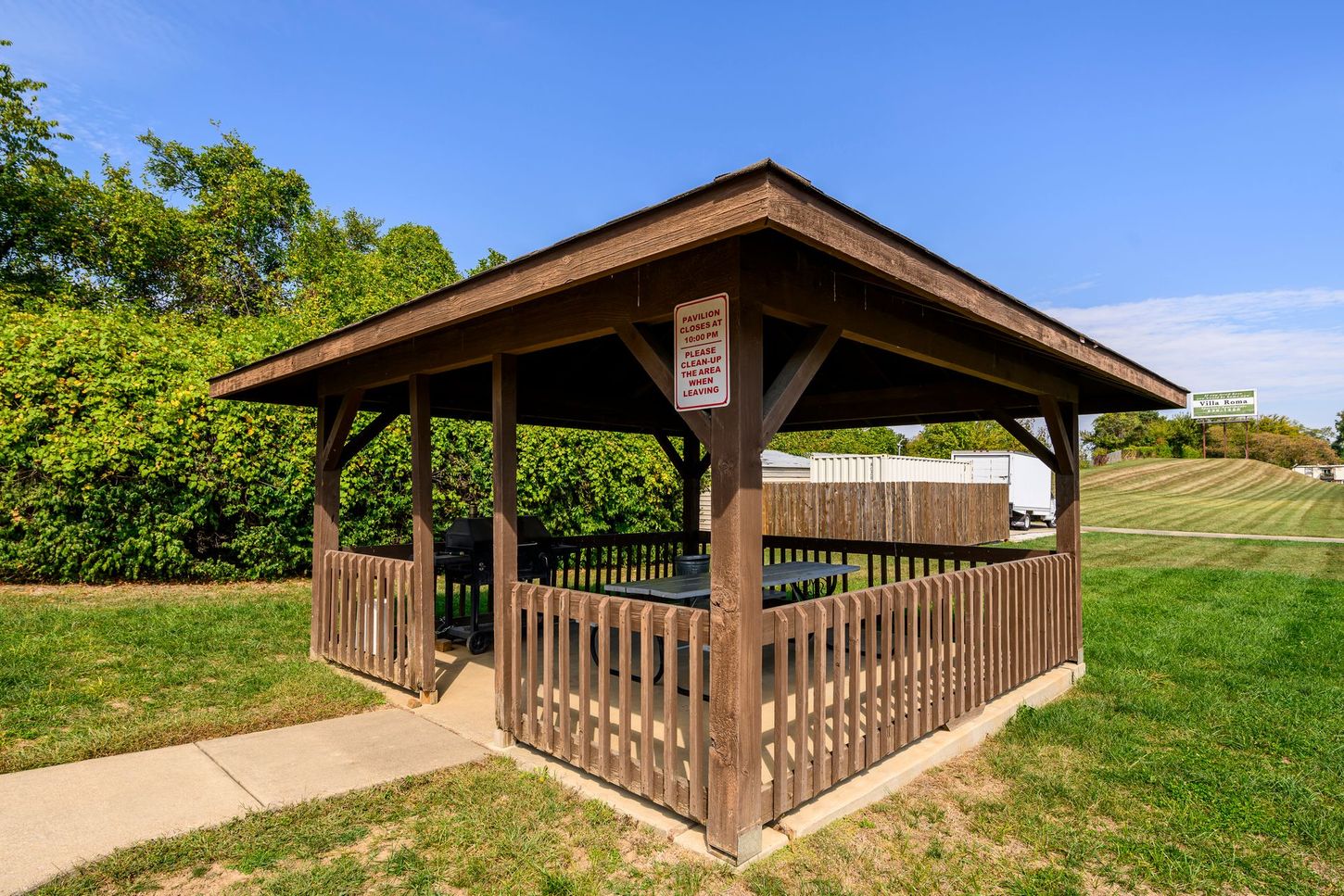 Brown gazebo with picnic table on a grassy area, surrounded by a low wooden fence. Blue sky in the background.
