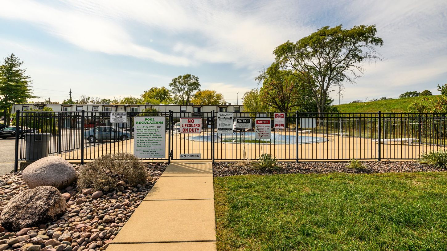 Fenced outdoor swimming pool with various posted signs and a gate entrance on a sunny day.