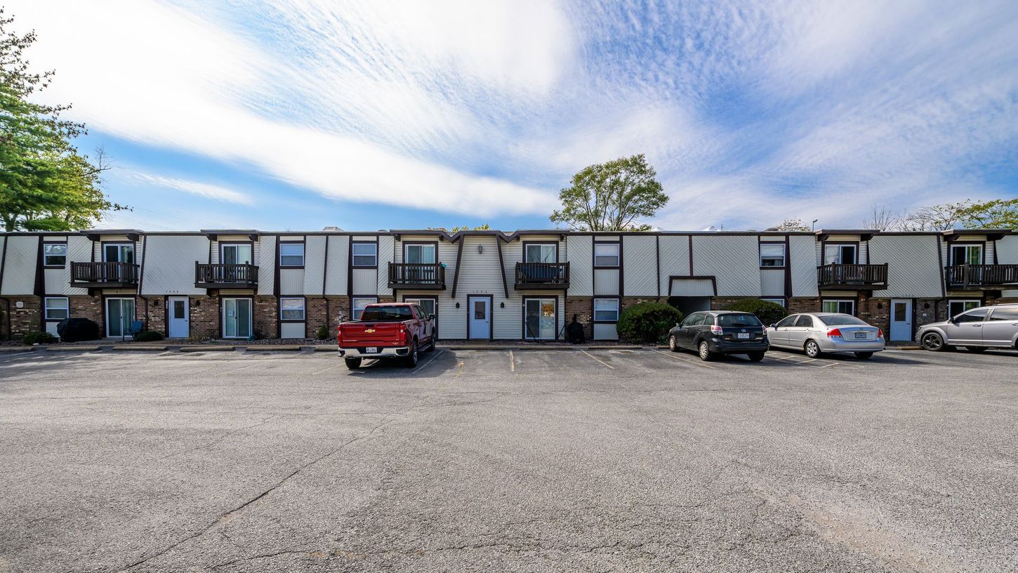 Row of apartments with small balconies and parked cars in front, under a blue sky with clouds.