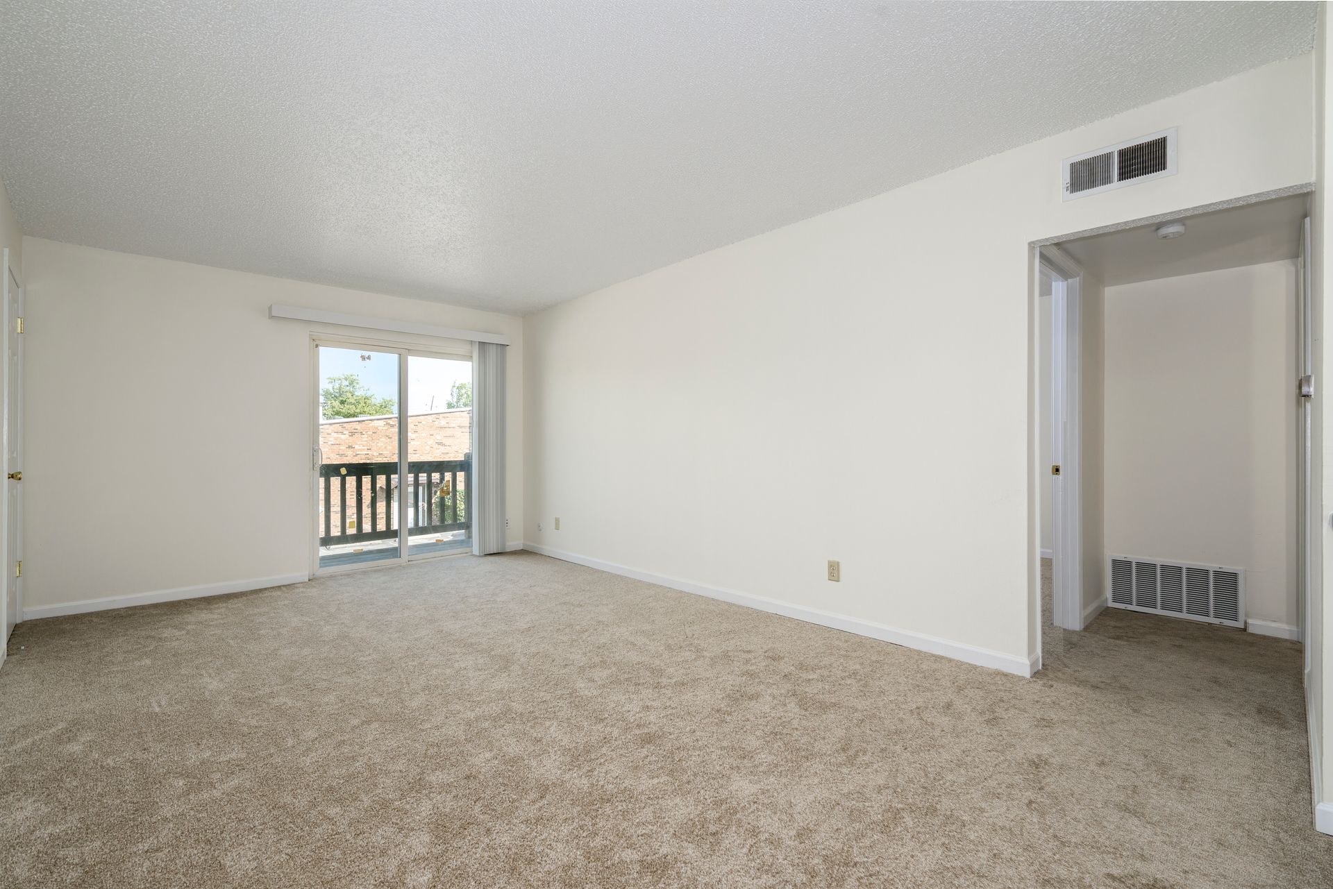 Empty carpeted room with sliding door to balcony and open doorway, white walls, and popcorn ceiling.