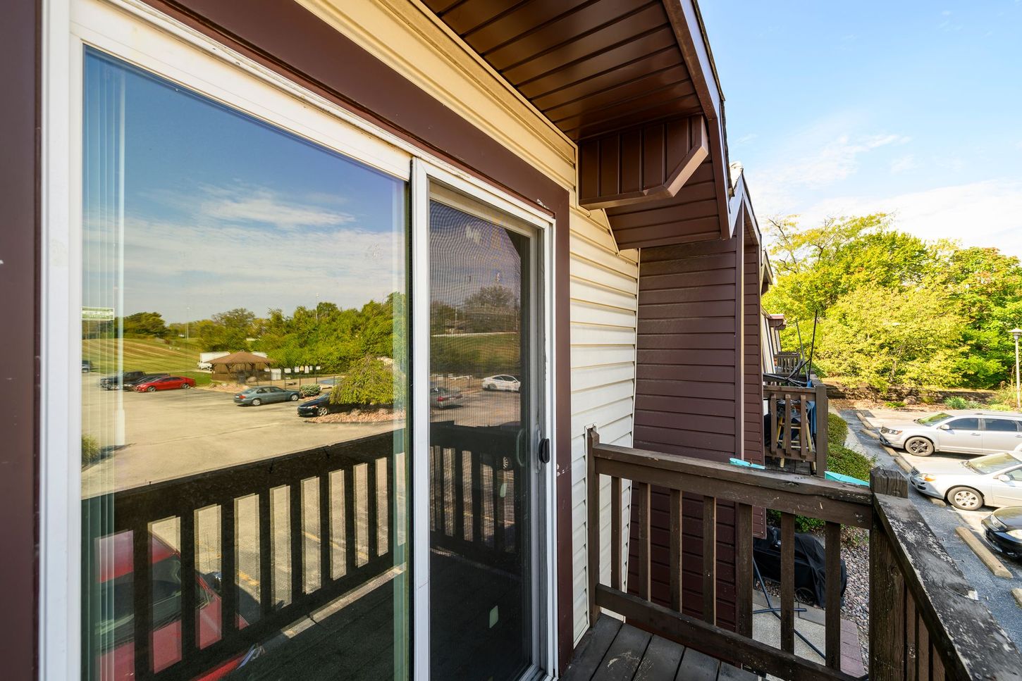 Balcony view: sliding glass door, wooden railing, parking lot visible, brown building exterior, sunny day.