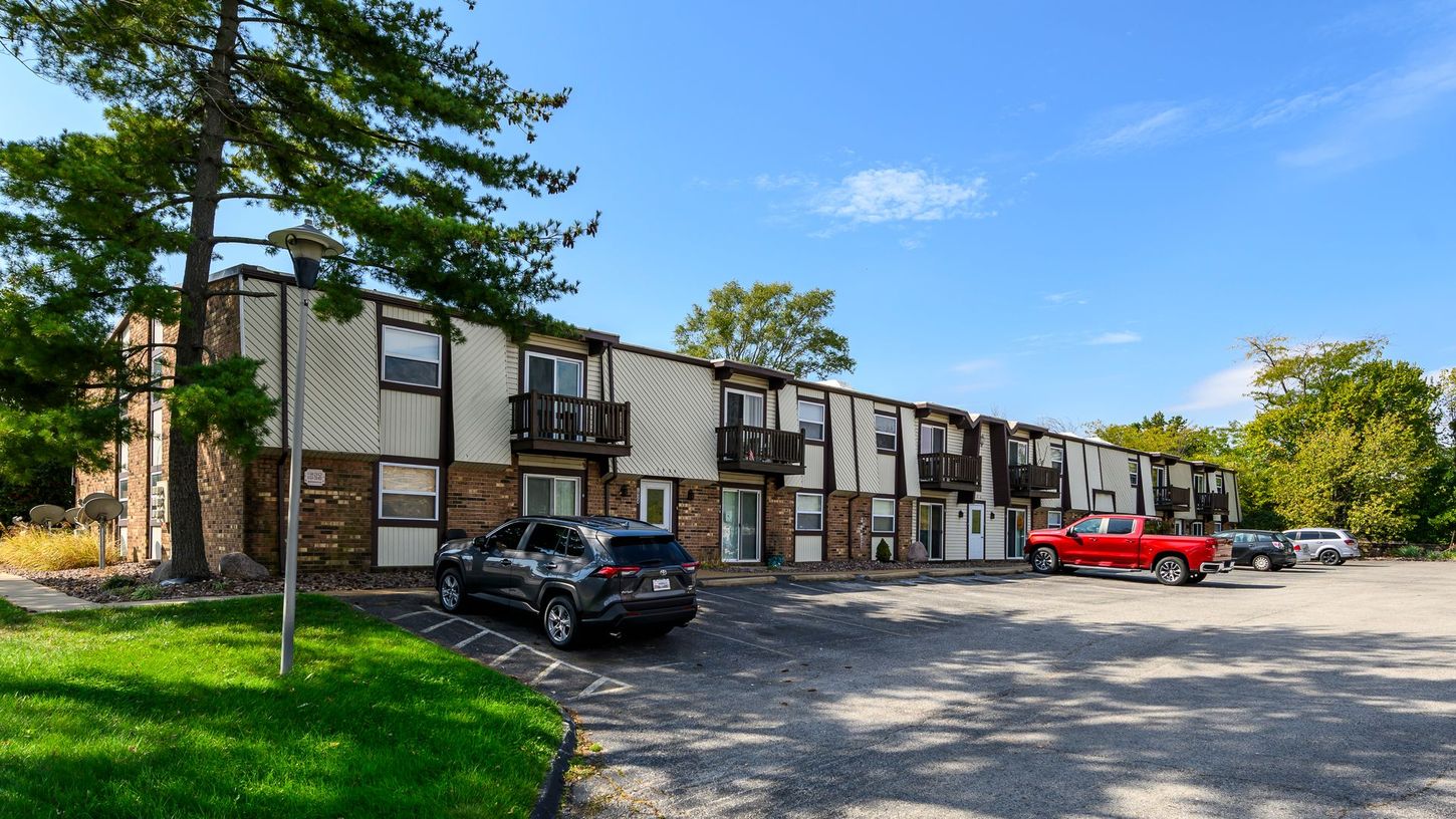 Apartment complex with brick and tan siding, cars parked in front, and green lawn under a blue sky.
