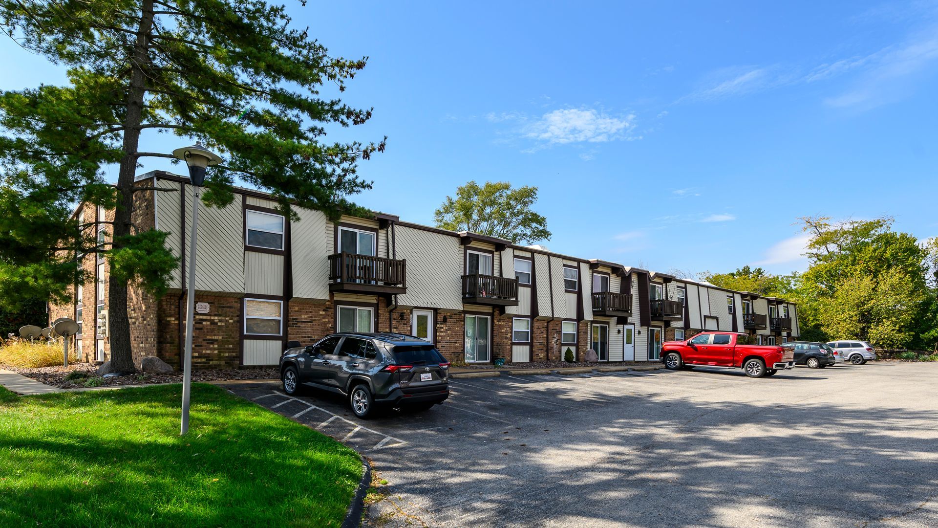 Apartment complex with brick and tan siding, cars parked in front, and green lawn under a blue sky.