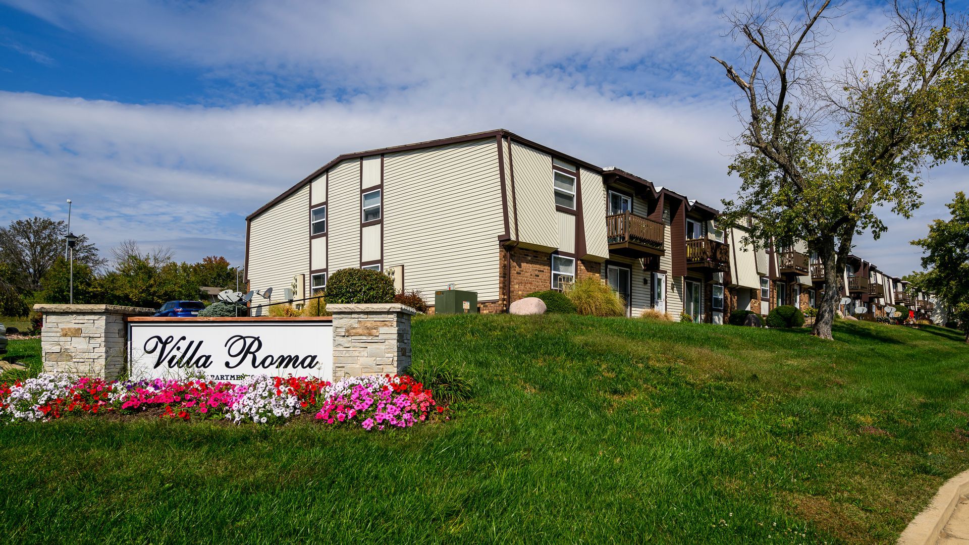 Villa Roma apartment sign in front of a tan two-story building on a grassy hill; flowers below the sign