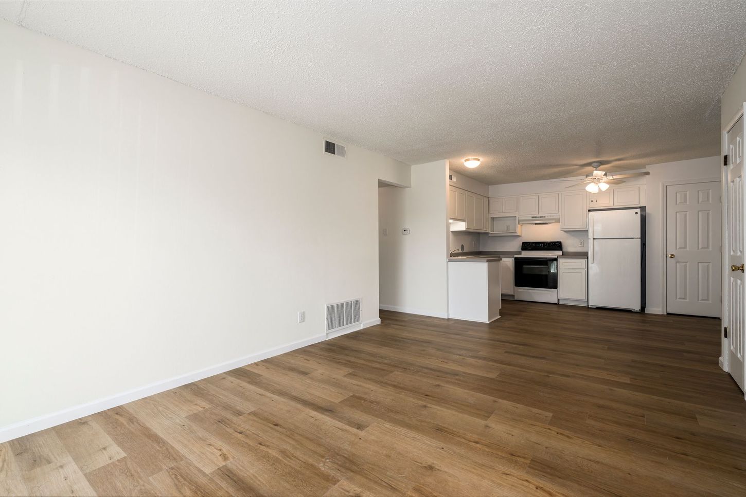 Empty apartment interior: white walls, wood floors, kitchen with white cabinets, and a refrigerator.