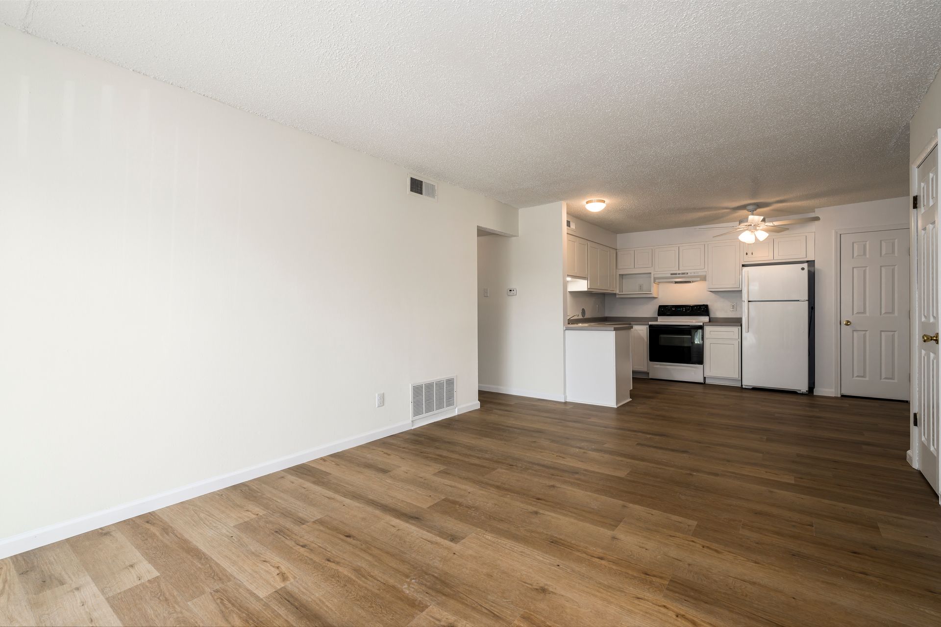 Empty apartment interior: white walls, wood floors, kitchen with white cabinets, and a refrigerator.