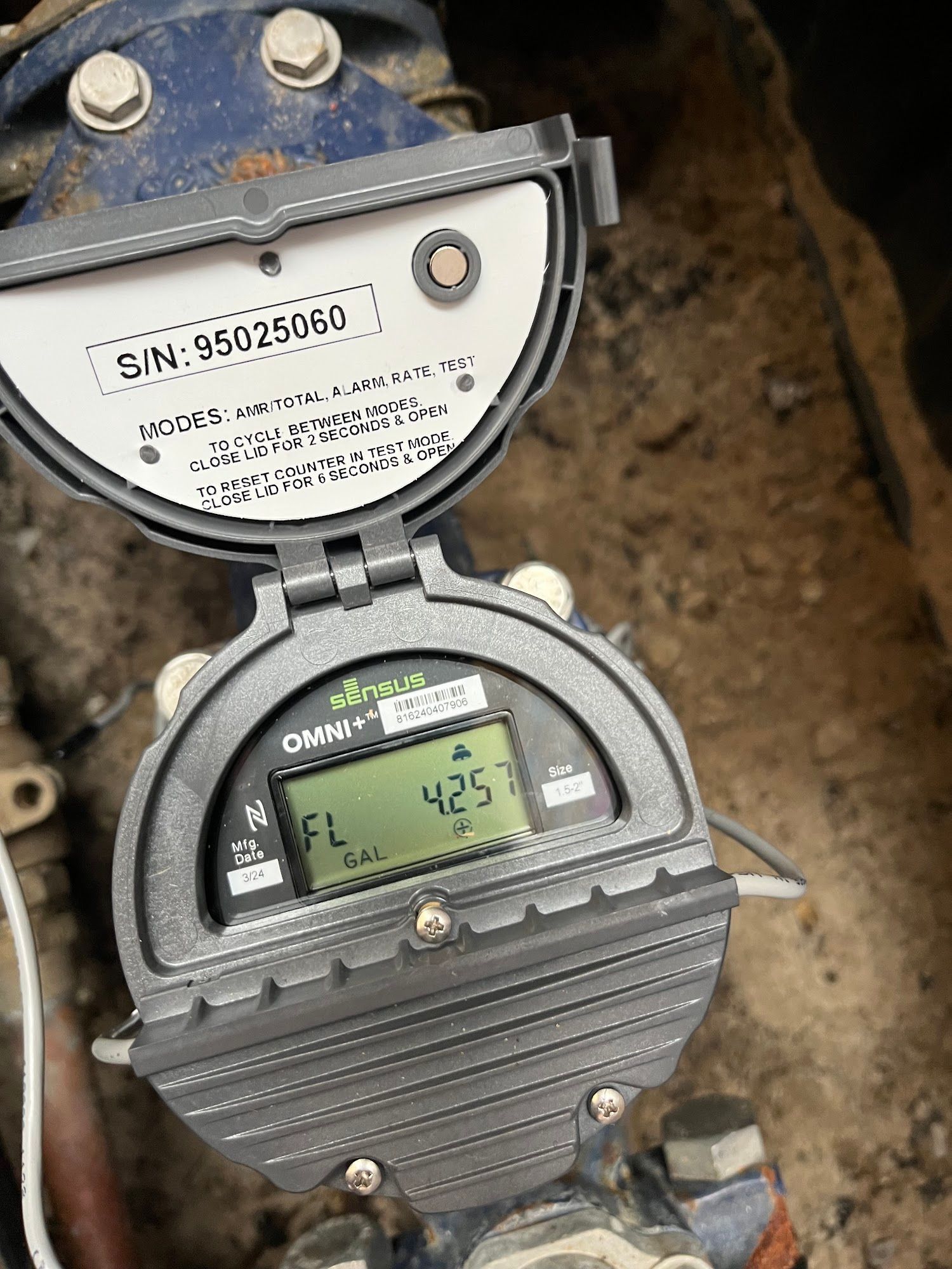 A water meter is sitting on the ground next to a pipe.