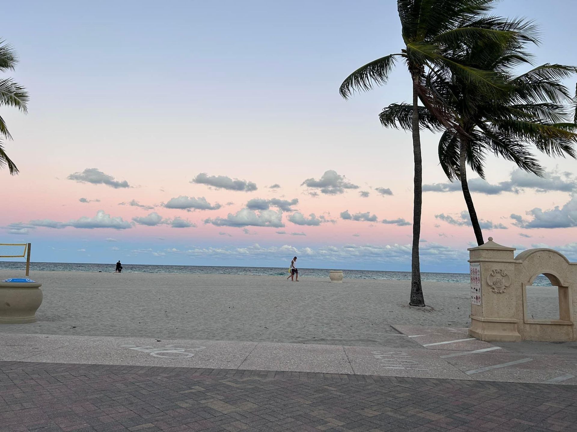 Beach at dusk with pink and blue sky, palm trees, and a person walking on the sand.