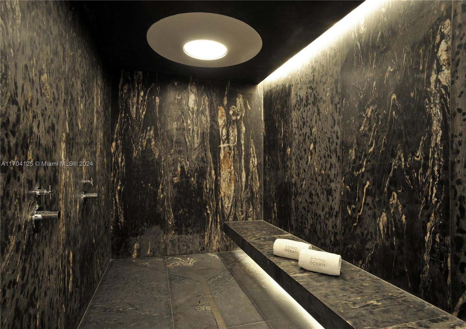 Dark stone spa shower with a built-in bench and towels, bright overhead light.