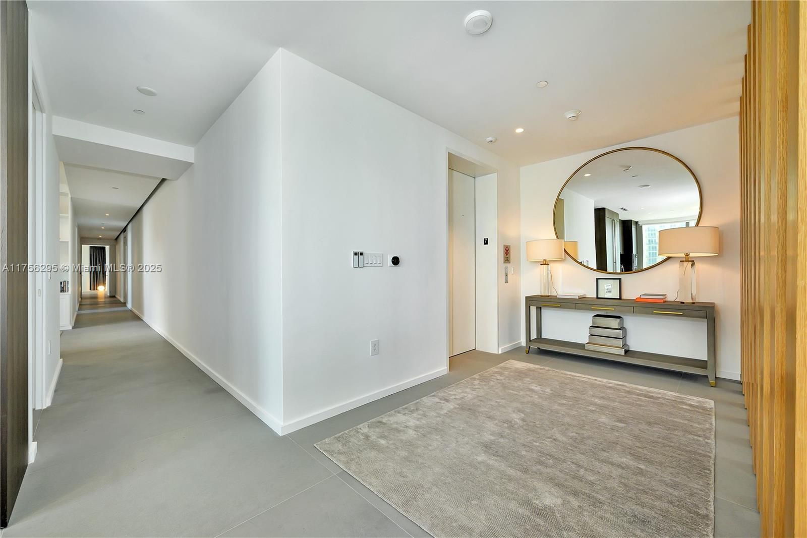 Long, white hallway with light gray flooring, leading to an entry with a rug, console table, mirror, and elevator.