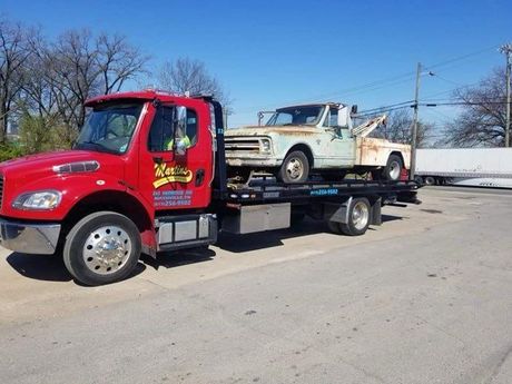Red tow truck carrying a weathered white pickup truck on a bright day.