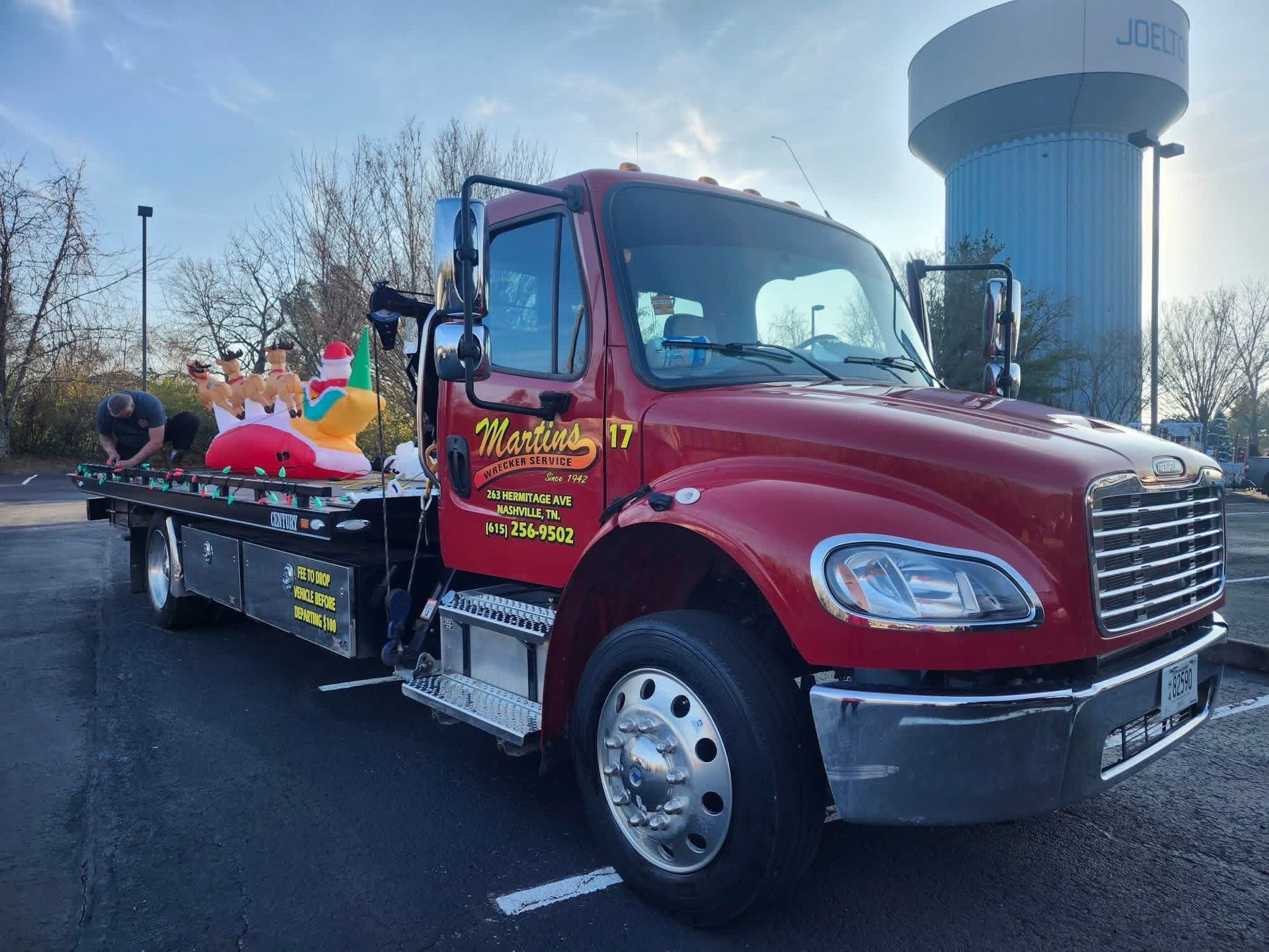 Red tow truck with toys on the bed, parked in front of a water tower.