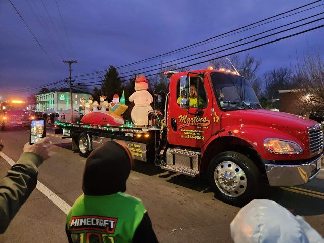 Red tow truck in a parade with Christmas lights and decorations, driven by a person.