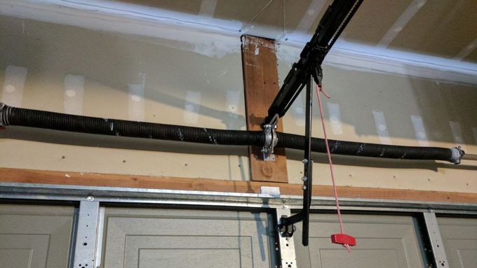 Garage door mechanism: spring, track, and opener components. Brown door frame, beige wall.