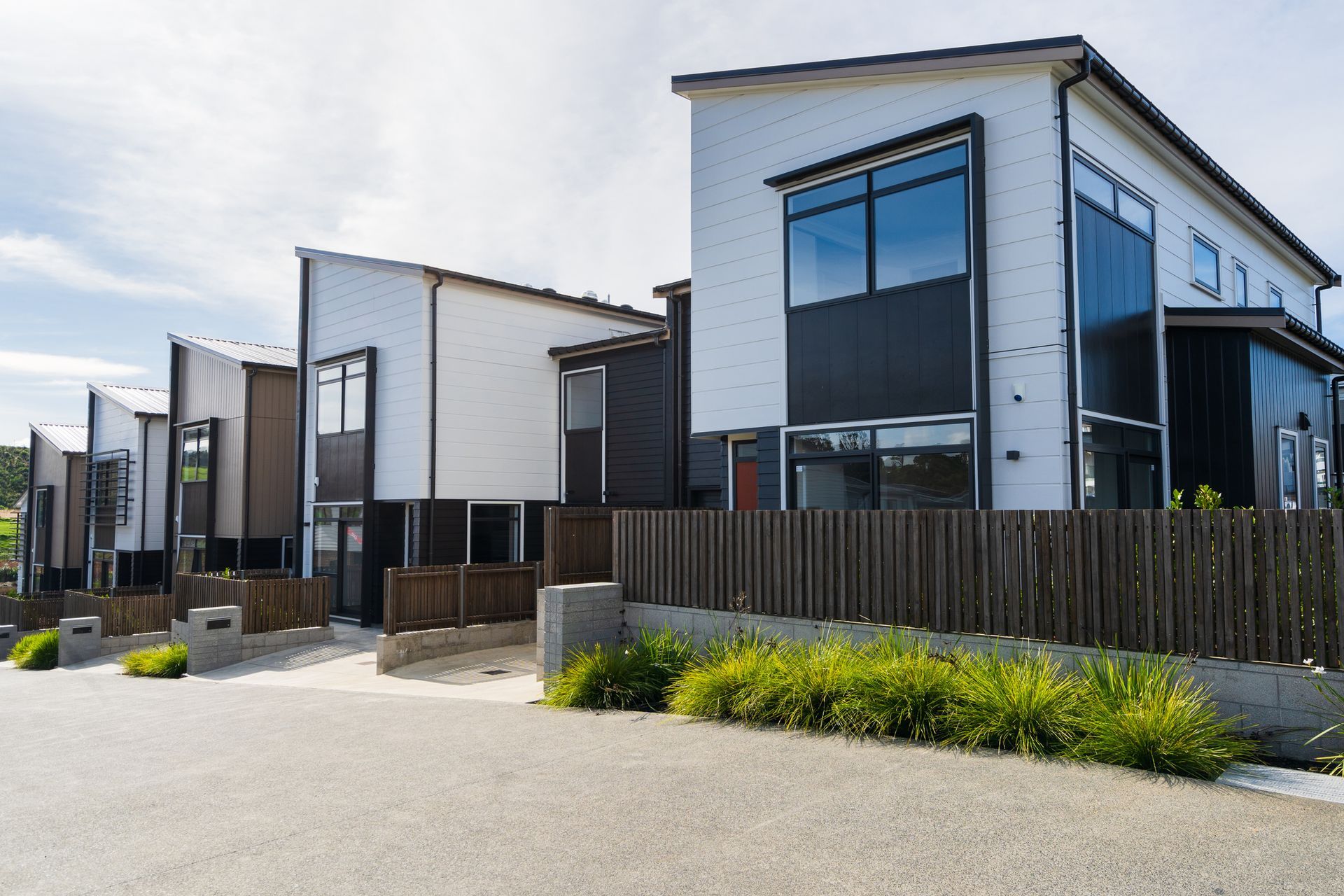 Modern white and dark-paneled townhouses behind a brown wooden fence under a sunny sky.