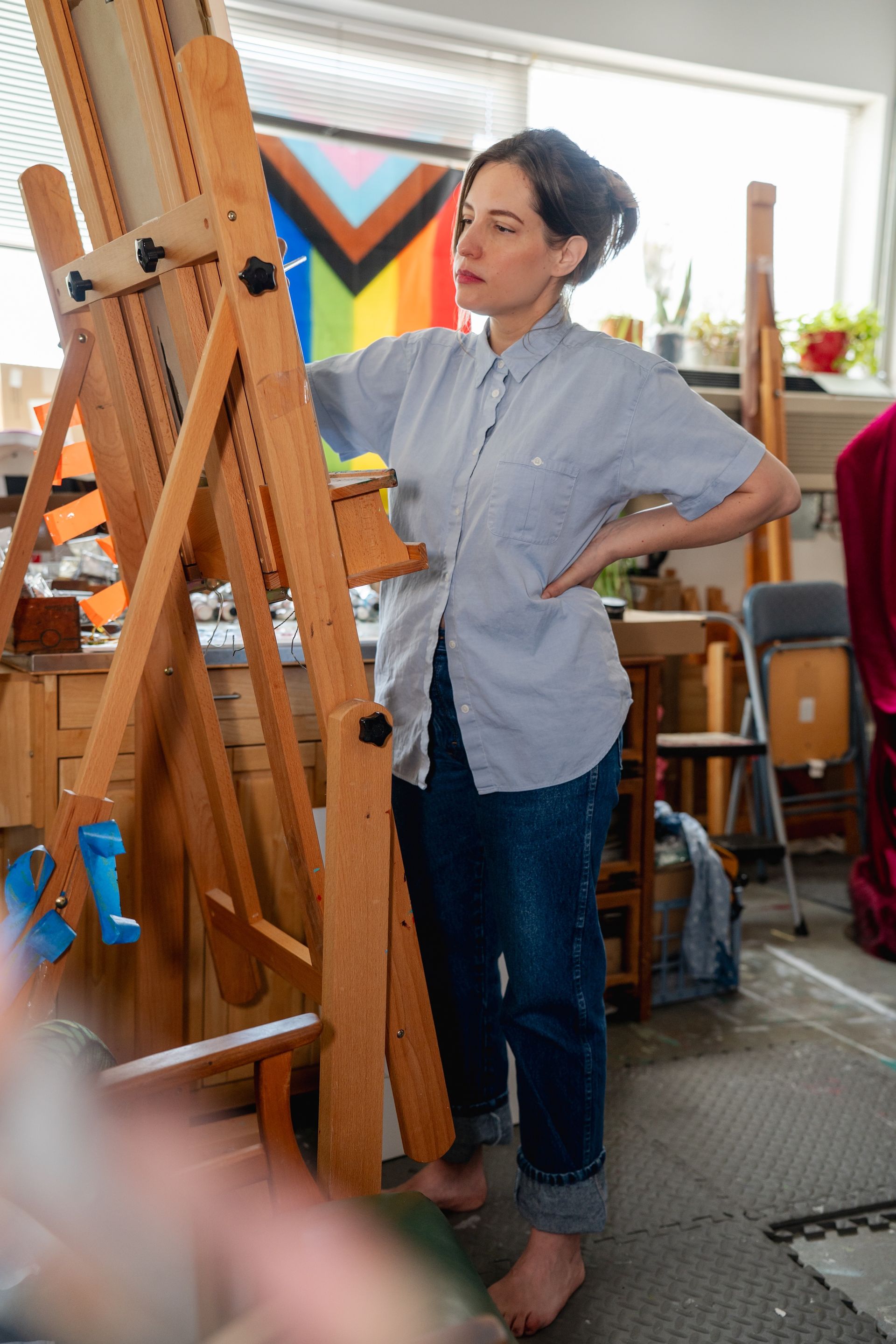 The artist in 2025, standing at her easel in her studio and wearing a blue button-down shirt.