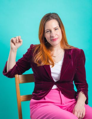 The artist in 2021, seated and smiling, wearing a velvet blazer and pink pants. 