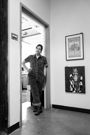 A black and white photo of the artist in 2025, standing in the doorway of her studio, wearing denim jumpsuit.
