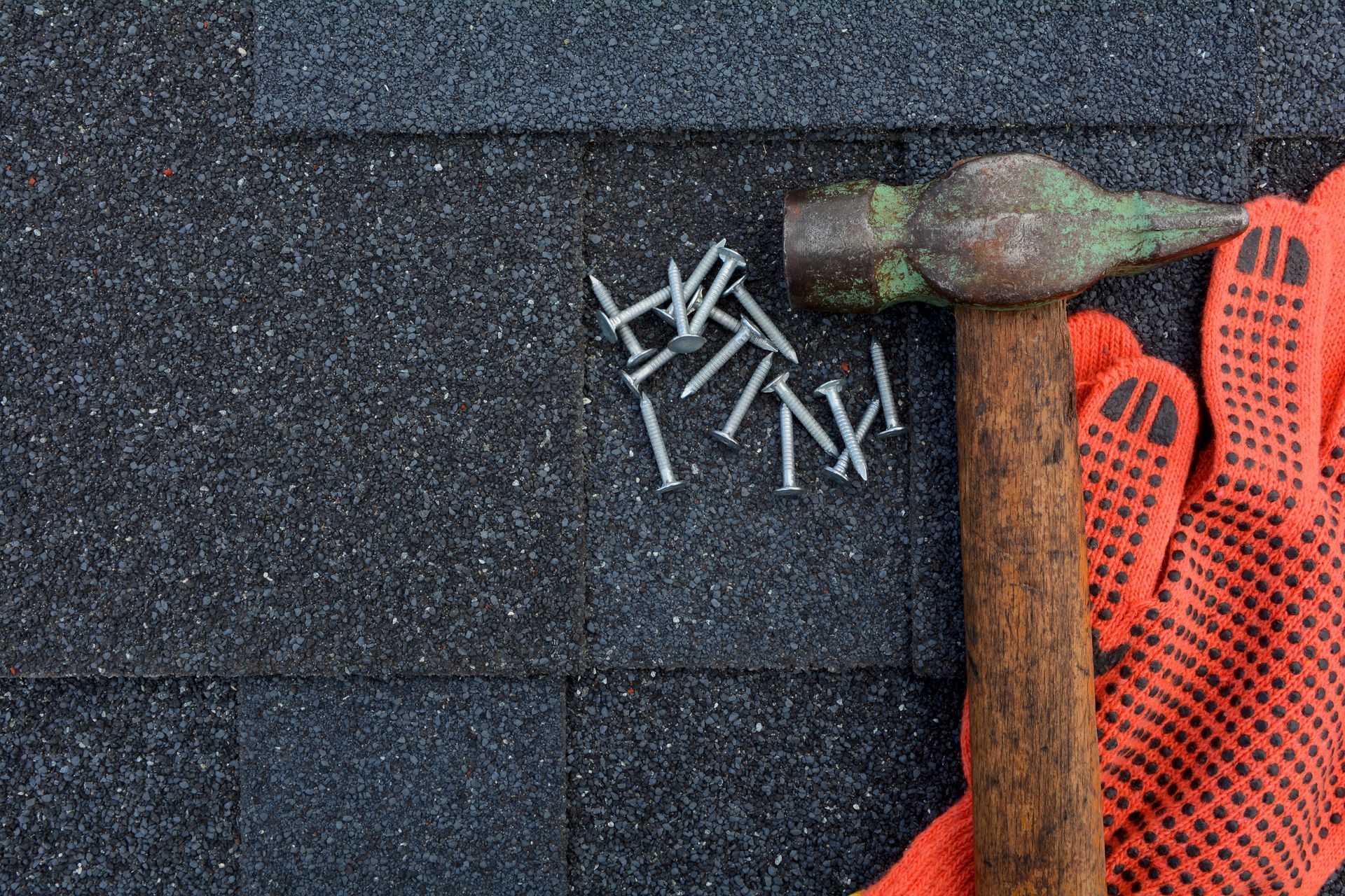 Hammer, nails, and orange work glove on dark gray roofing shingles.