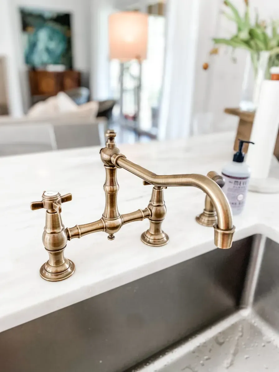 Antique-style bronze kitchen faucet on a white countertop with a stainless steel sink in a well-lit kitchen.
