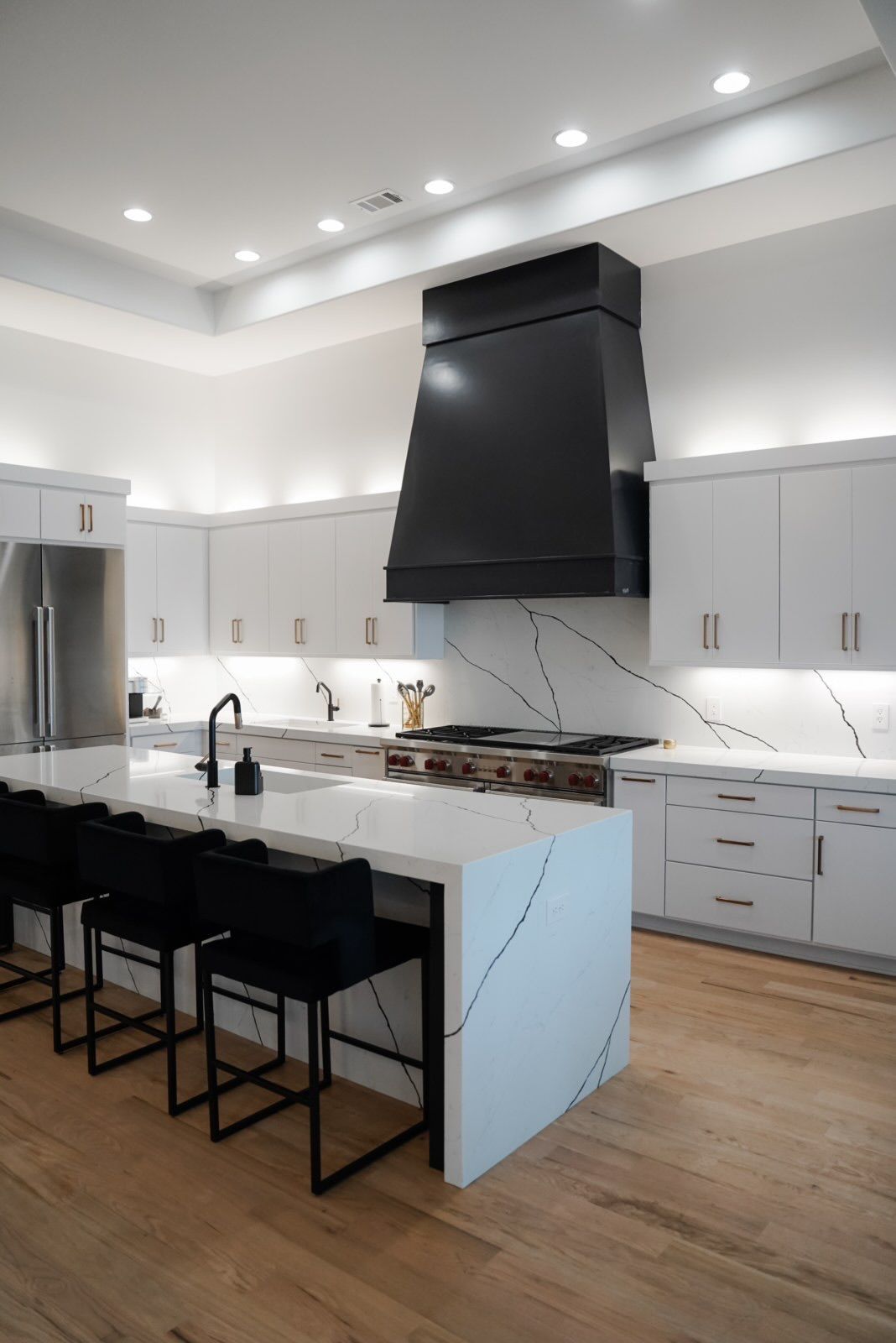 Modern white kitchen with black range hood and island.  Quartz countertops, wooden floor, and black bar stools.