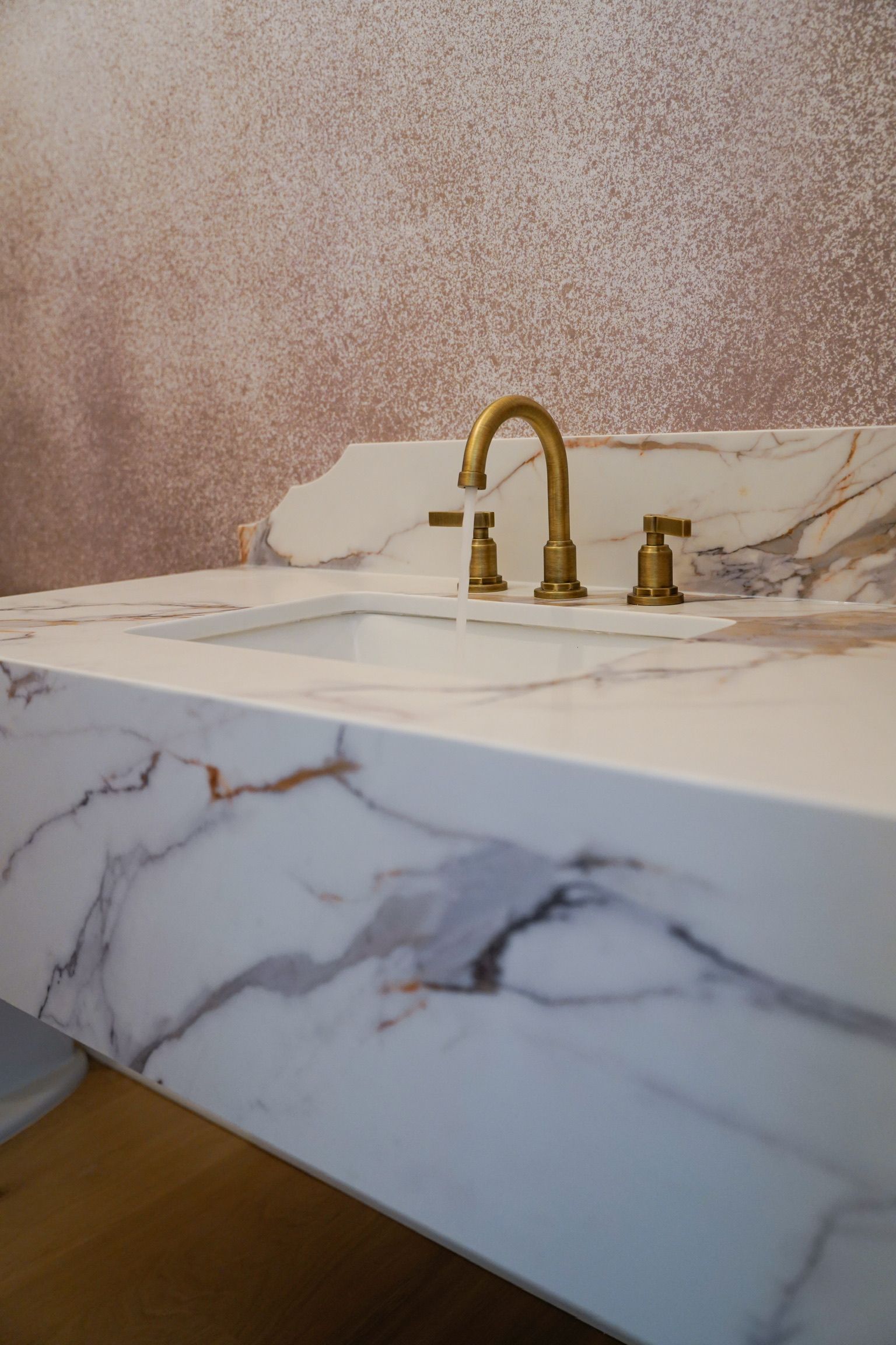 White marble countertop with gold faucet and basin; textured pink wall in background.