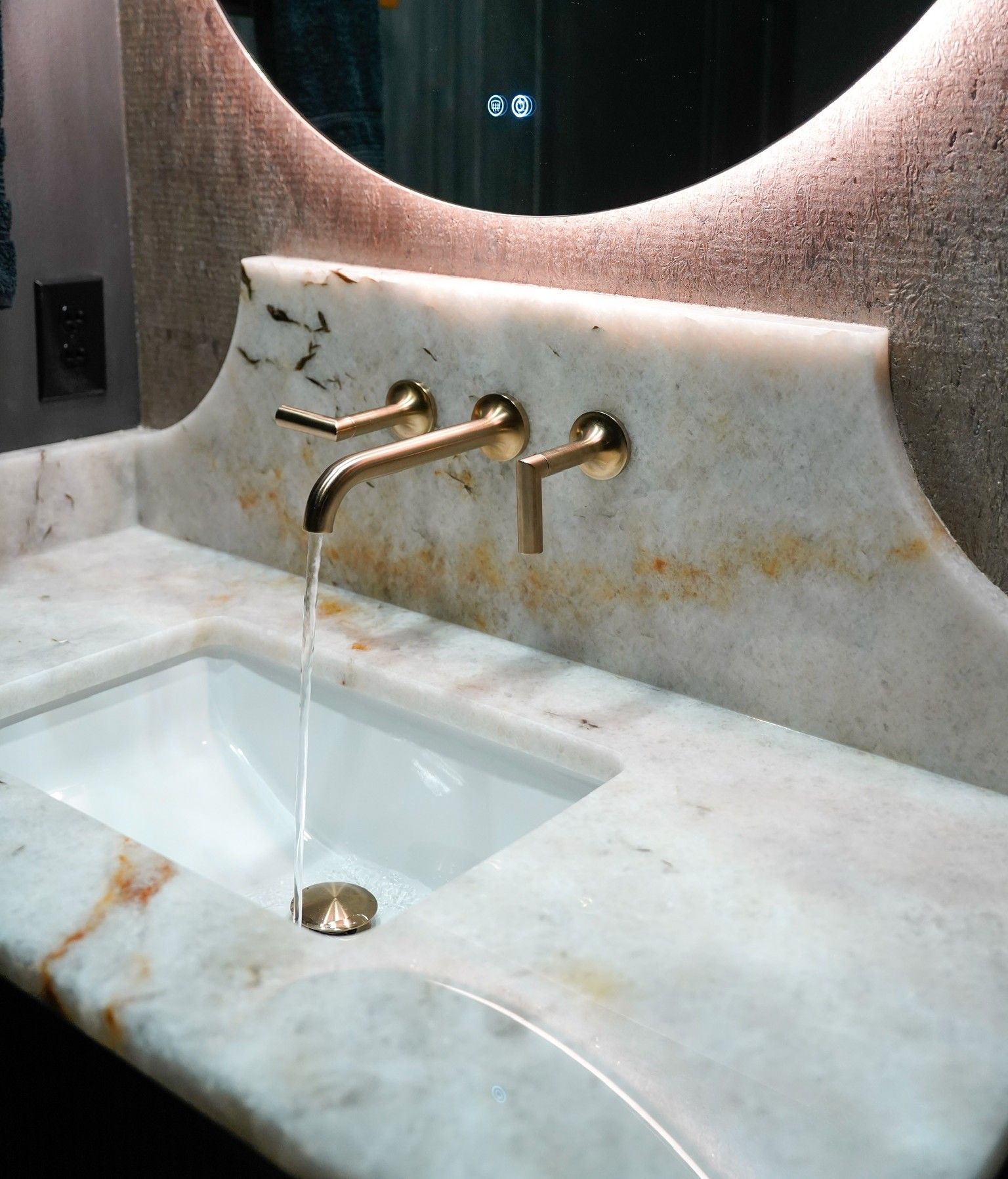 Bathroom sink with gold faucet, marble countertop, and oval mirror. Water streams into the white rectangular basin.