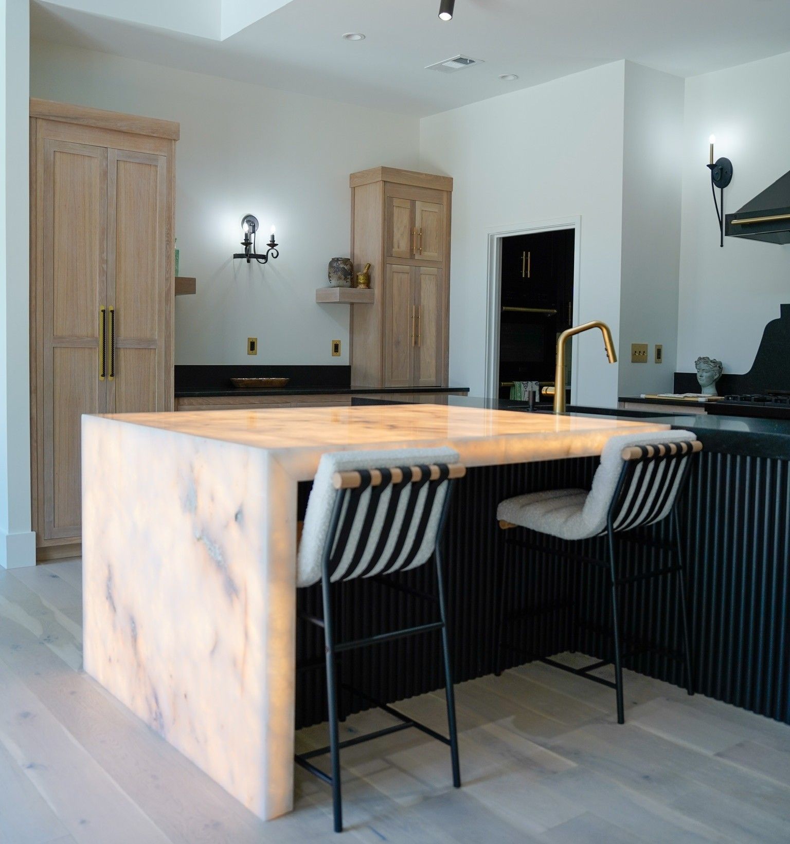 Kitchen with illuminated island, two bar stools, light wood cabinets, and black accents.