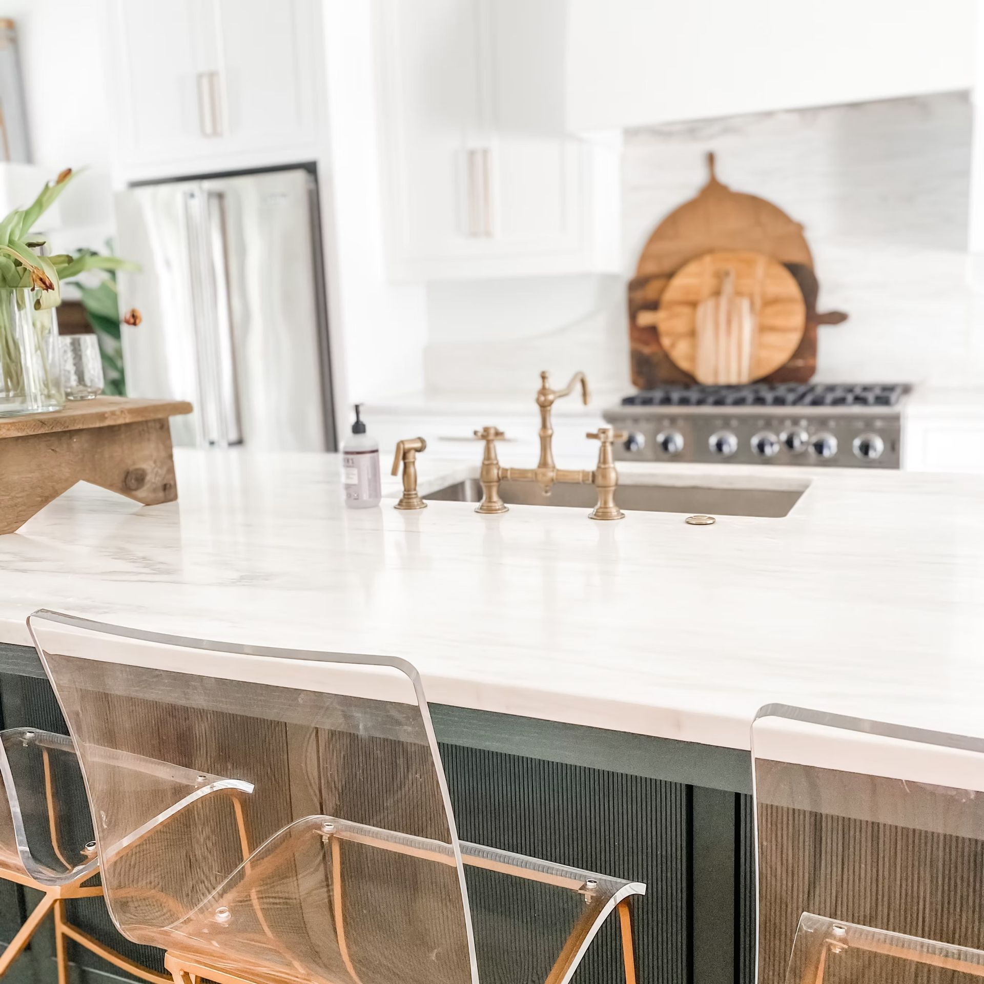 Modern kitchen with clear acrylic chairs, marble countertop, gold faucet, and wooden cutting boards.