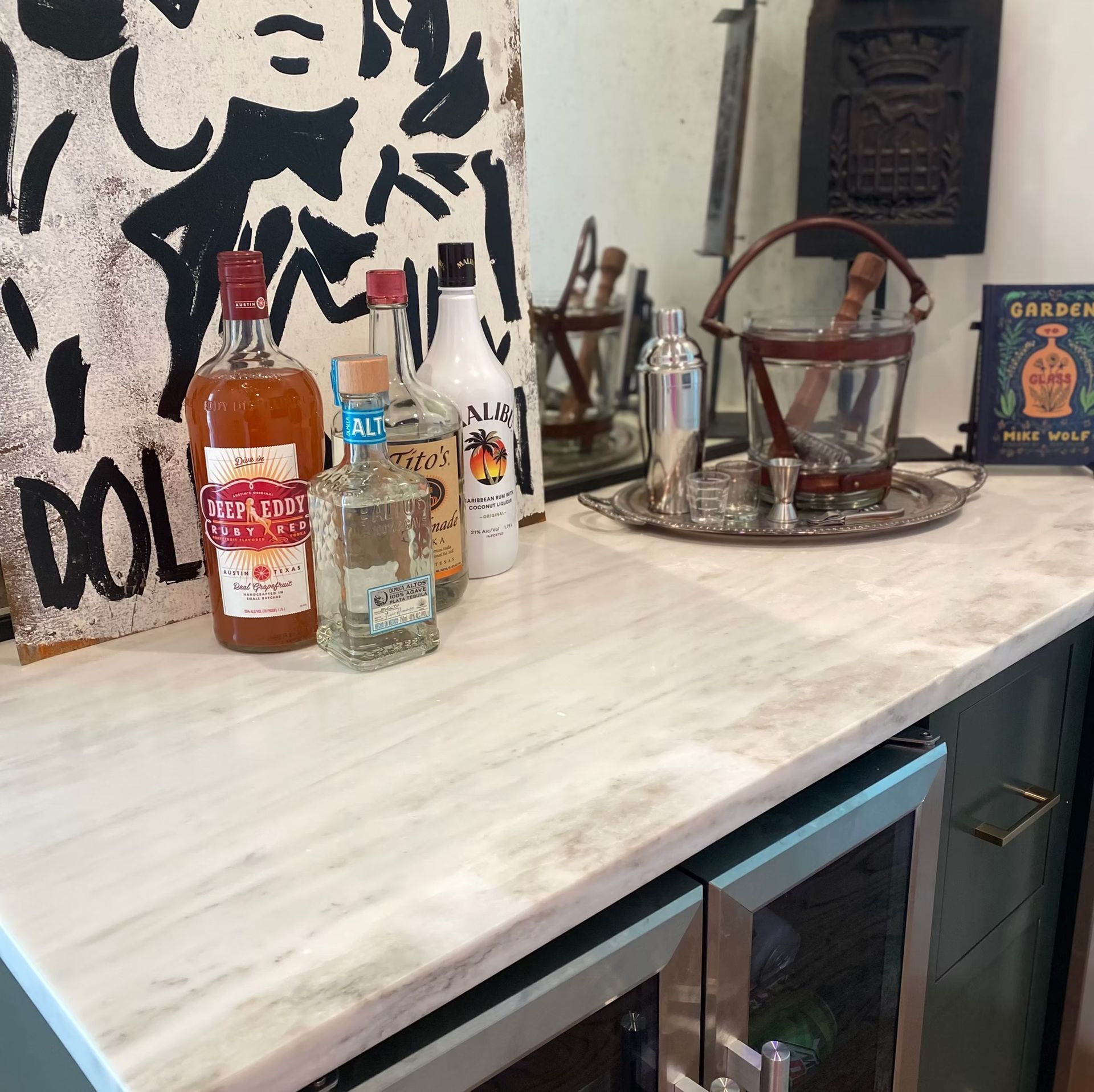 A home bar setup. Bottles of liquor, shaker, ice bucket, and sign on marble countertop.