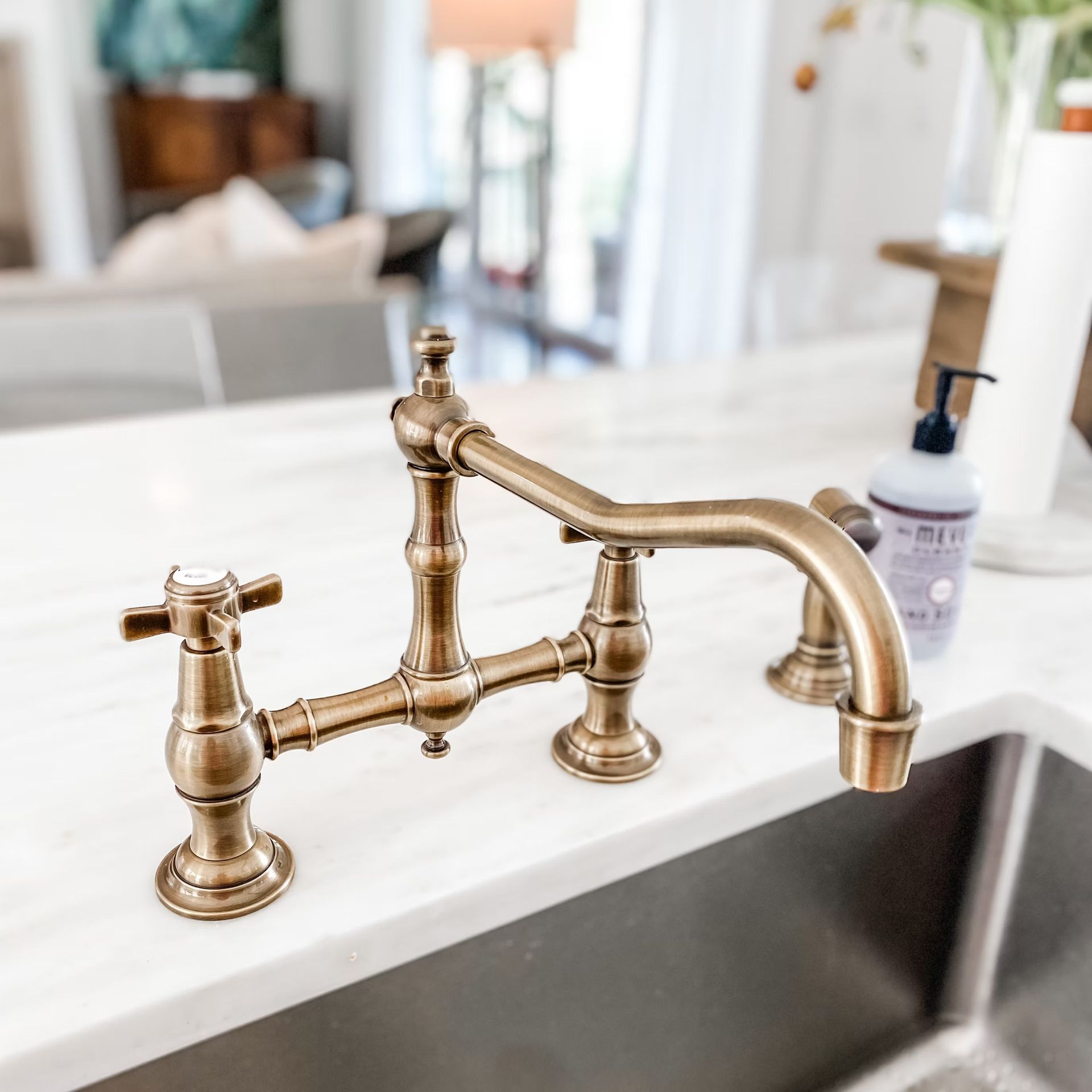 Bronze kitchen faucet on a white countertop over a stainless steel sink, with a soft-focus background.