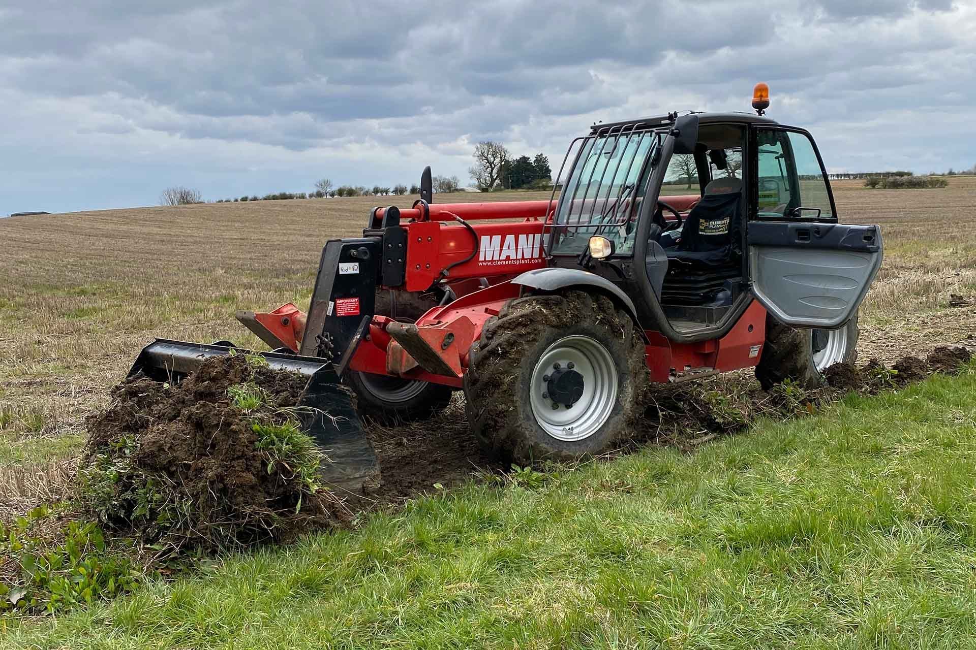 A DHS Plant Hire red Manitou telehandler driving through a field .