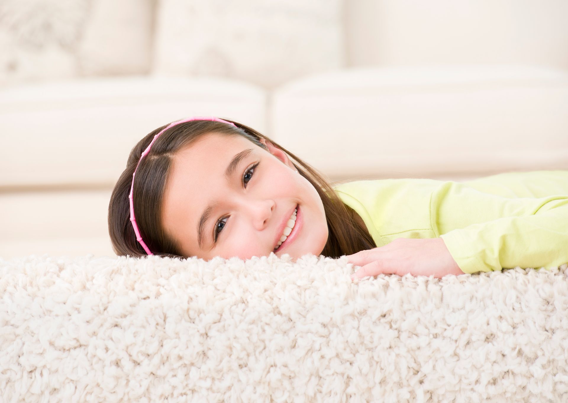 Girl with brown hair and pink headband smiles on a plush white carpet.