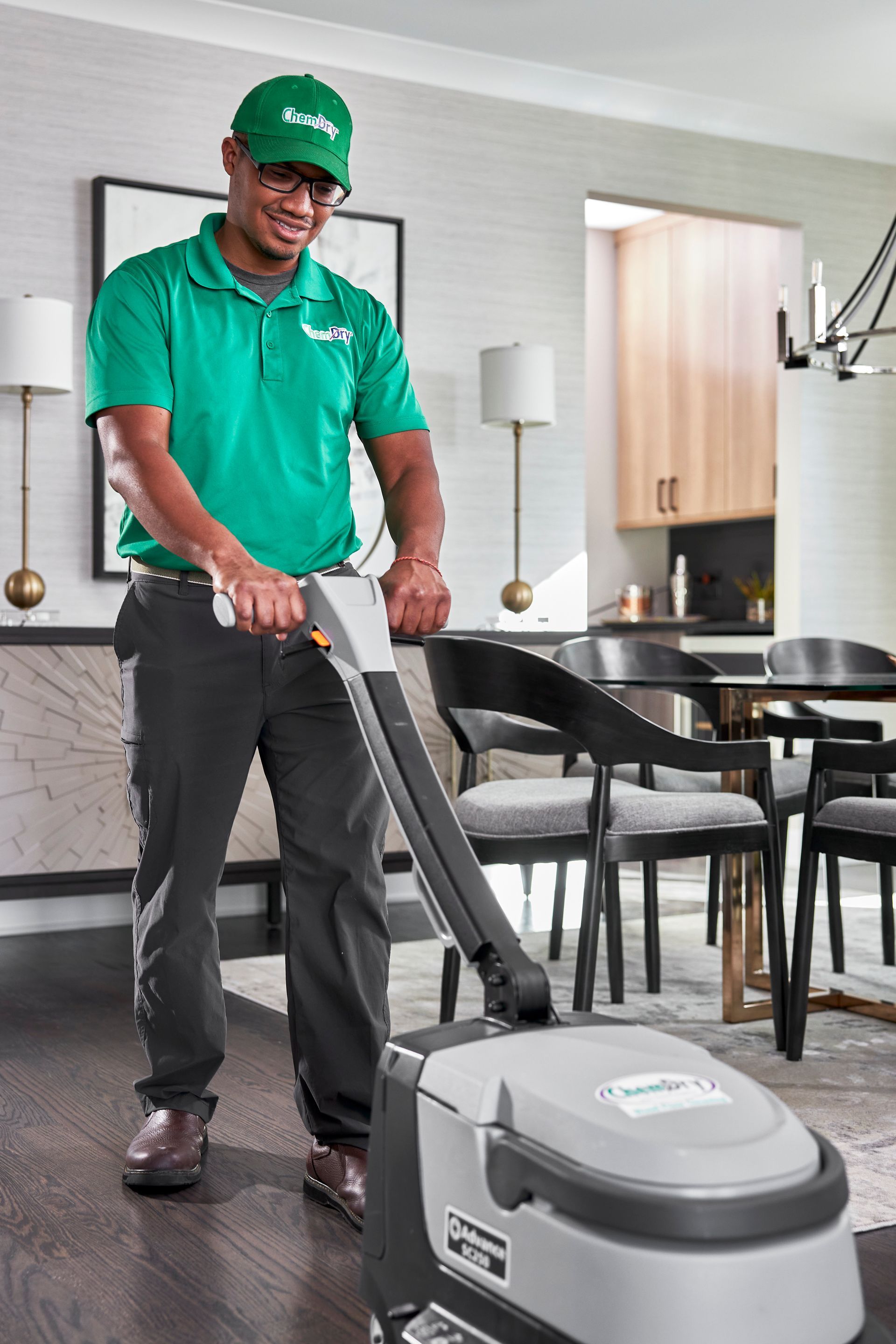 Man in green shirt and hat operating a floor cleaner in a living room.