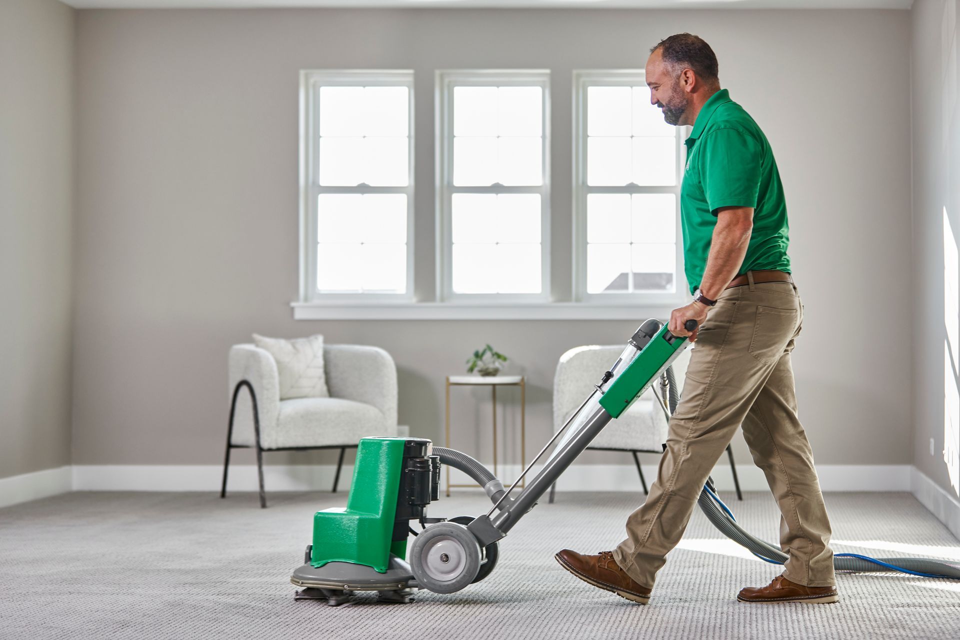 Man using carpet cleaner in a room with windows. He wears a green shirt and tan pants.