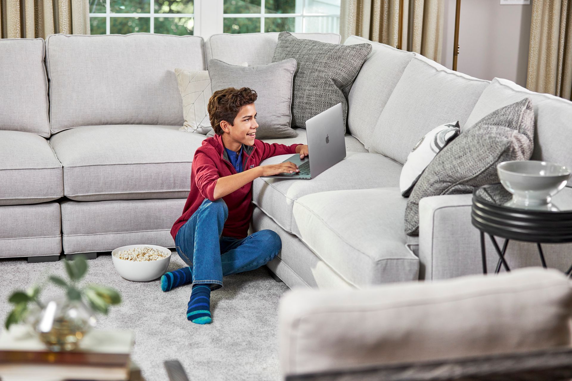 Person using a laptop while sitting on a light gray sectional couch, with popcorn bowl.