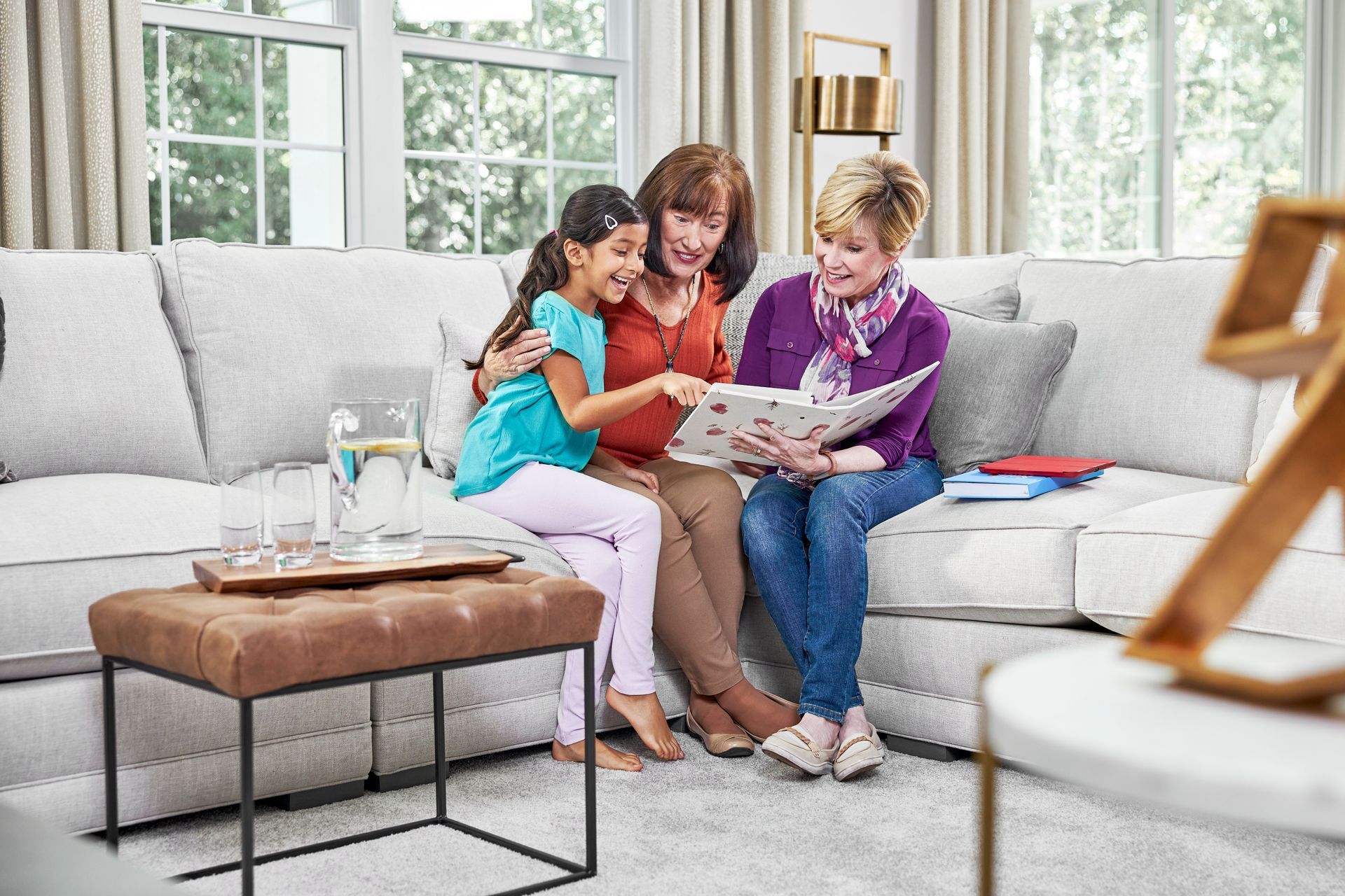 Three people looking at photo album on a couch.  Girl, woman, and older woman smiling, living room setting.