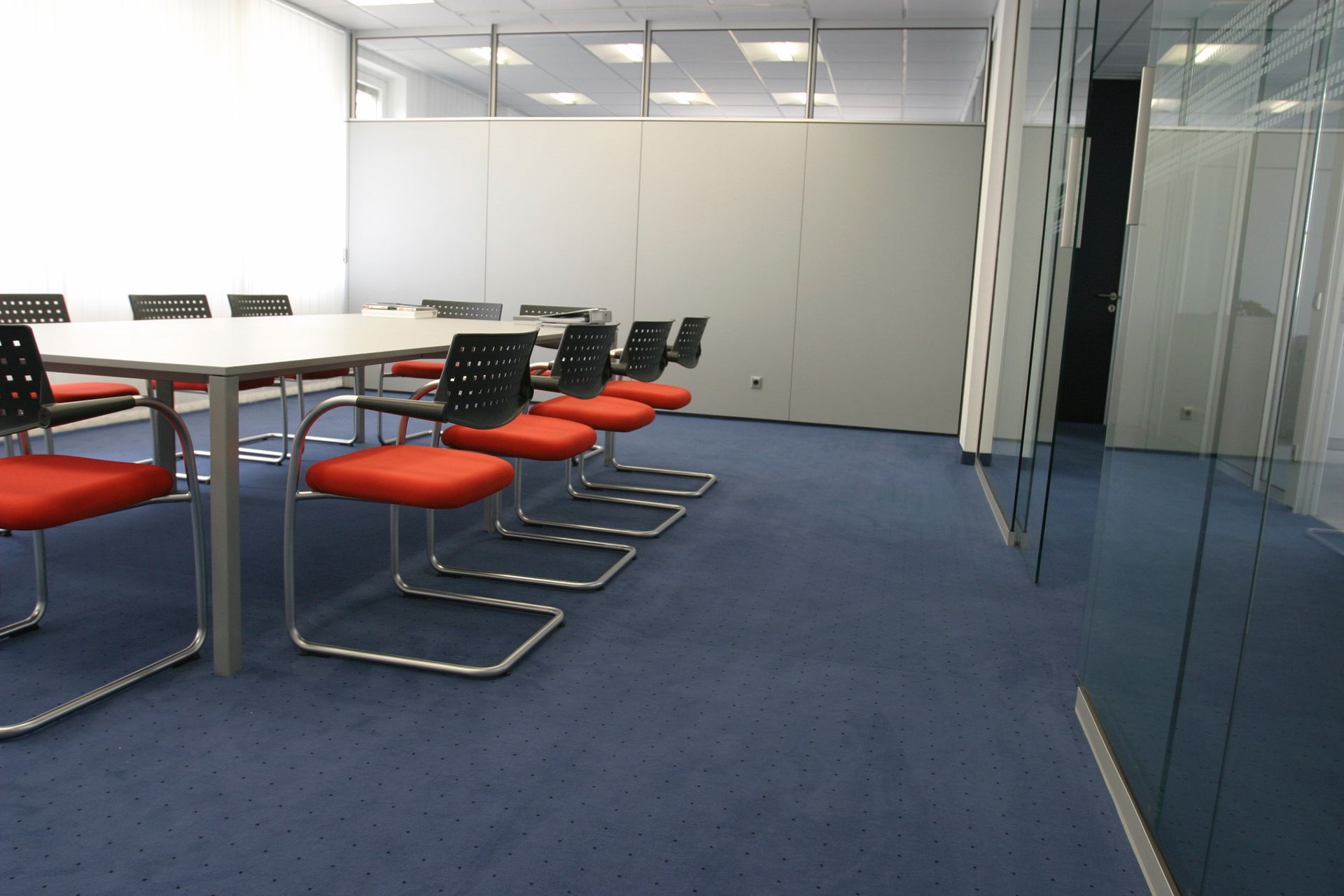 Conference room with a long white table, red chairs, and blue carpet.