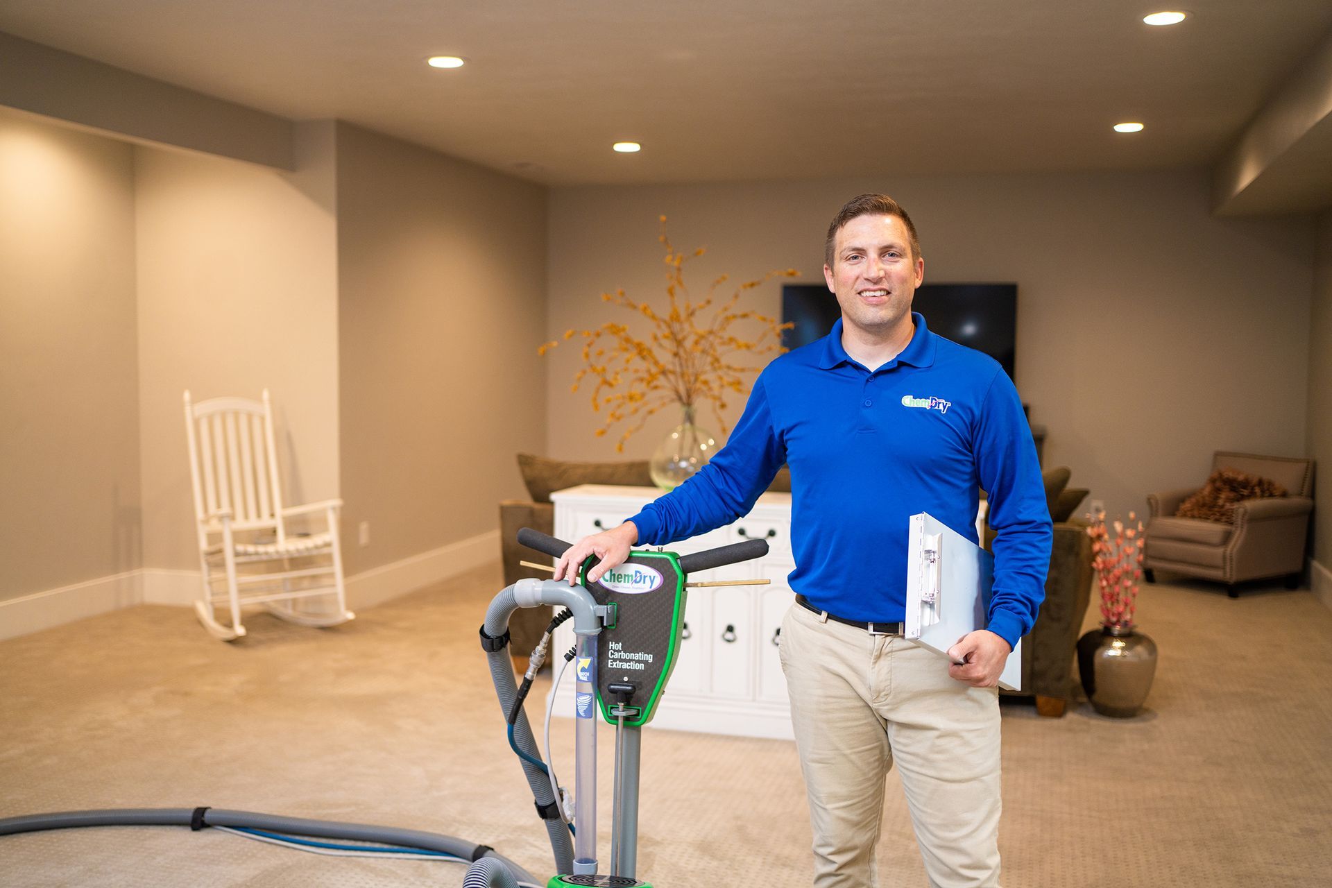 Man in blue shirt, tan pants, holding clipboard, standing with carpet cleaning machine in a living room.