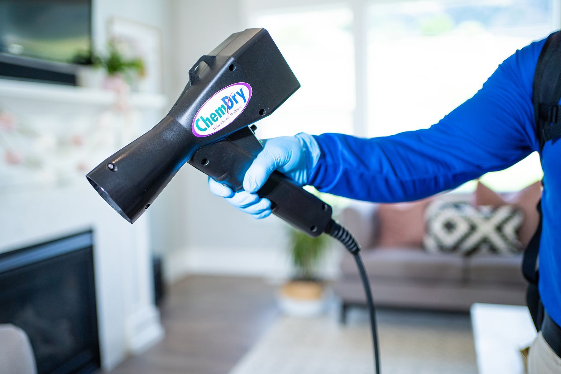 Person in blue shirt and gloves holding Chem-Dry sanitizing device indoors, near furniture.
